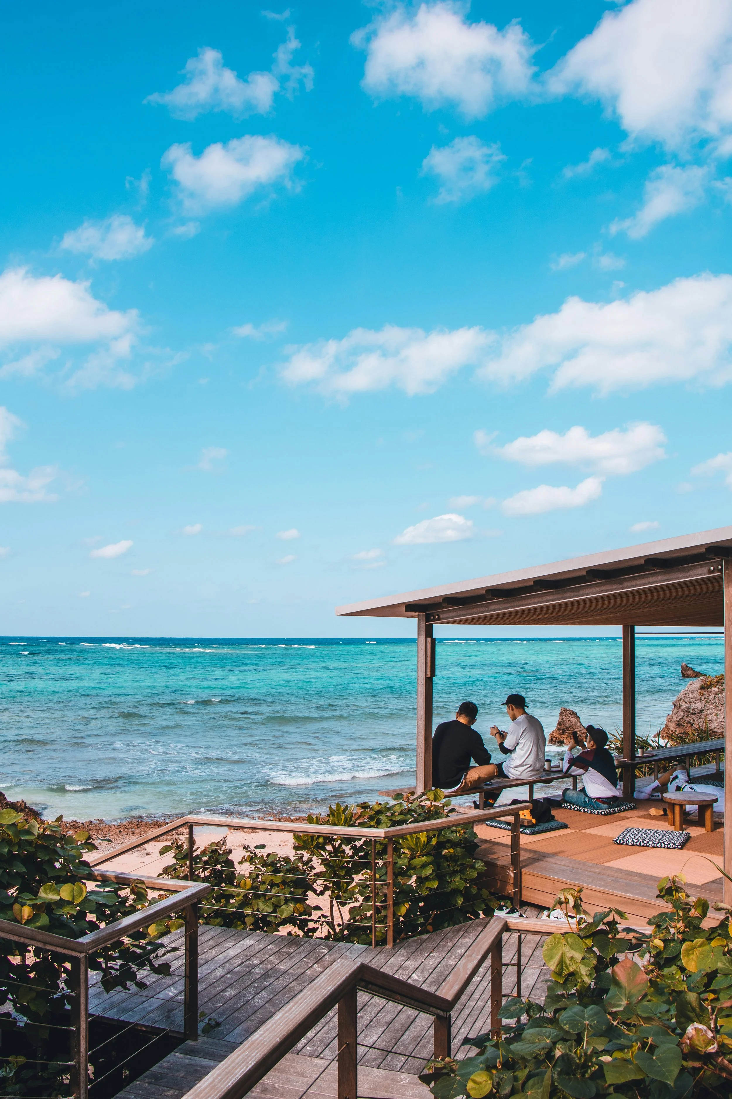 People sitting on a wooden deck with cushions under a roof on the beach, overlooking the ocean and sky with scattered clouds.