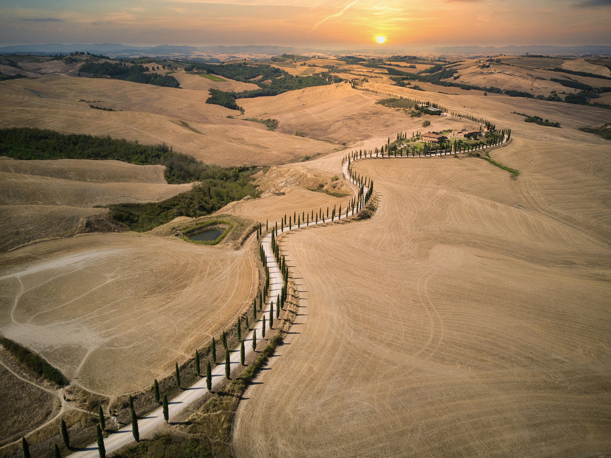 A winding dirt road lined with tall cypress trees across a landscape of arid, rolling hills at sunset in Tuscany, Italy.