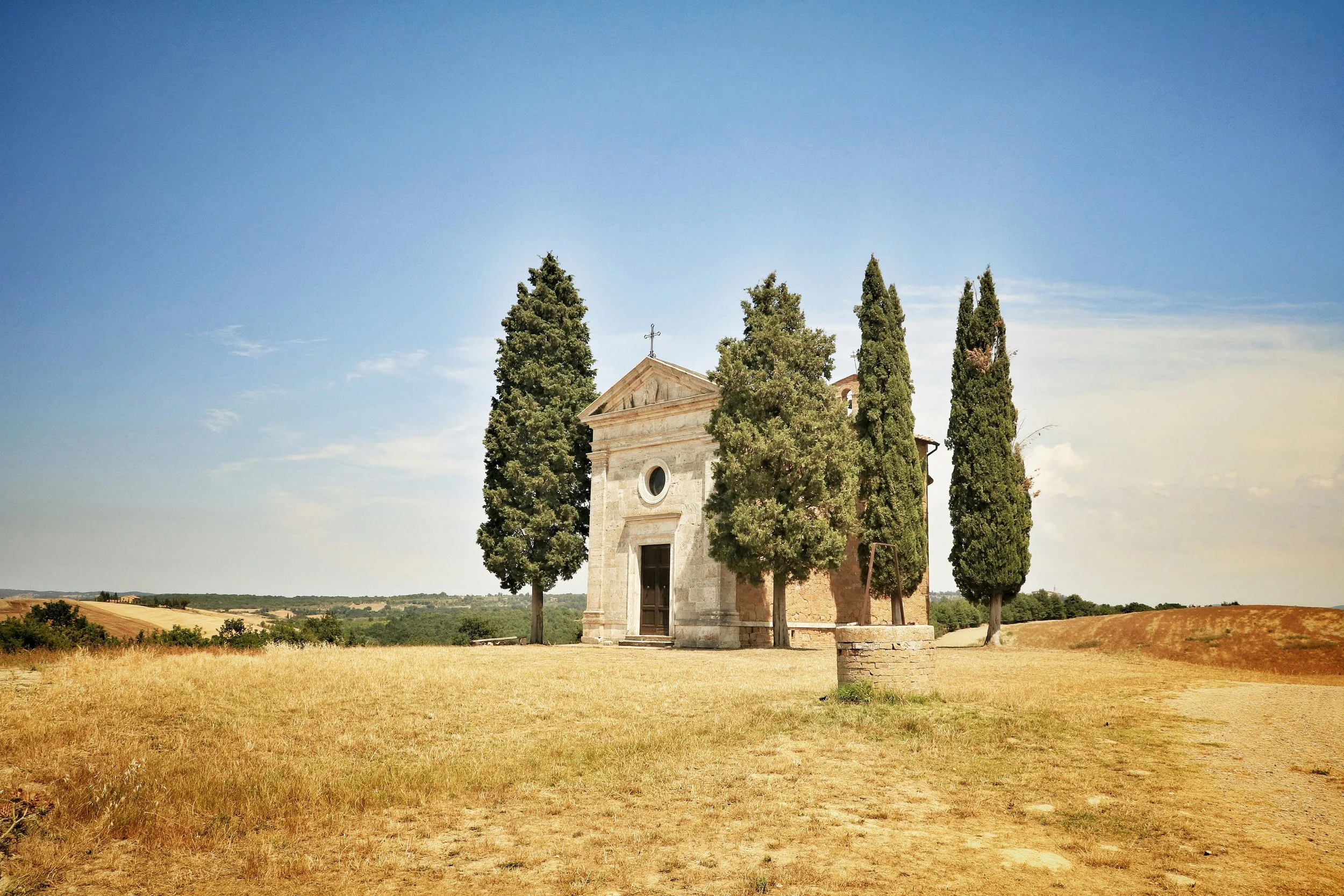 Small stone chapel surrounded by tall cypress trees on a grassy field under a blue sky.