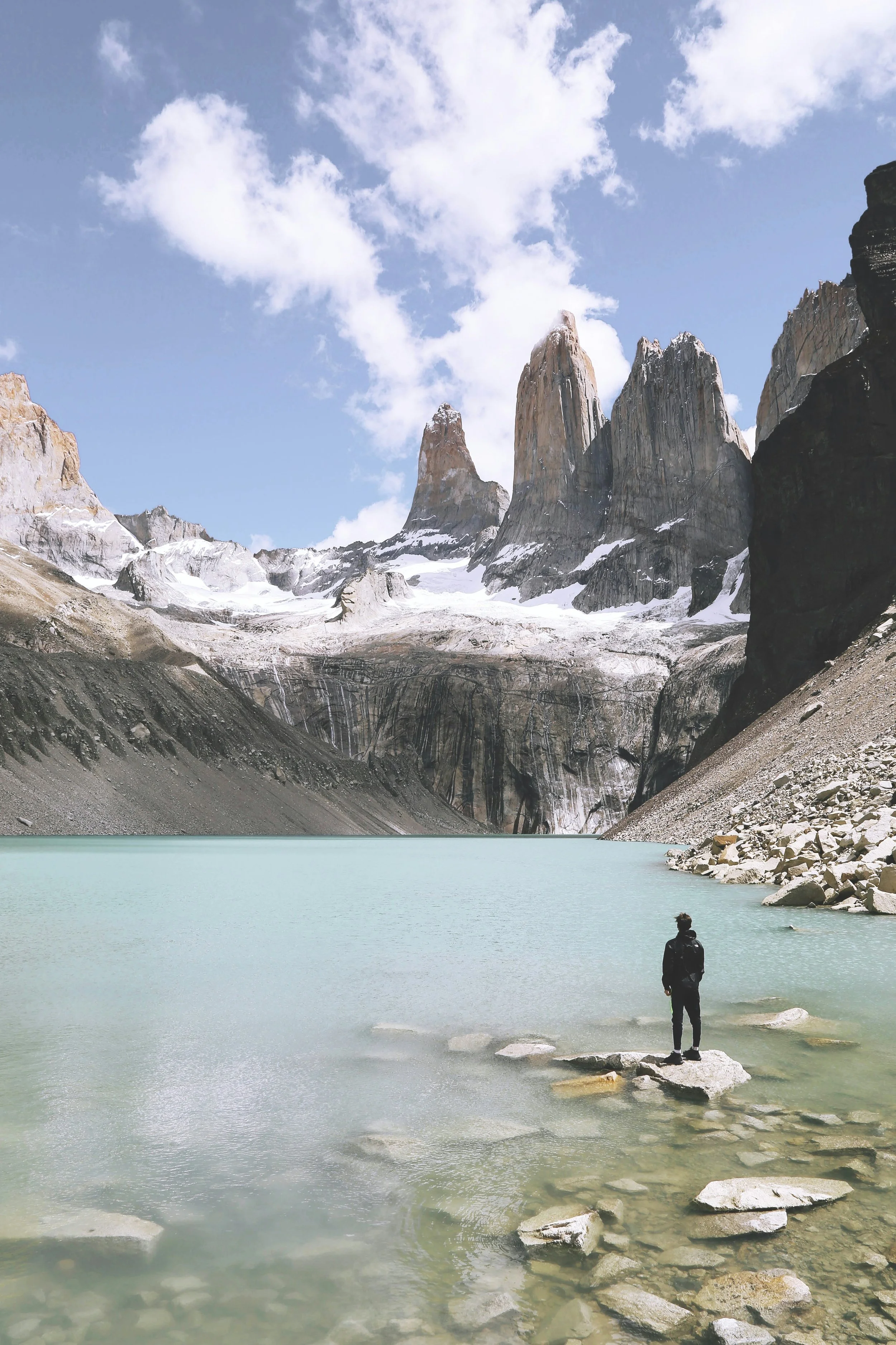 A person standing on a rock by a glacier-fed lake surrounded by mountains and snow-capped peaks.
