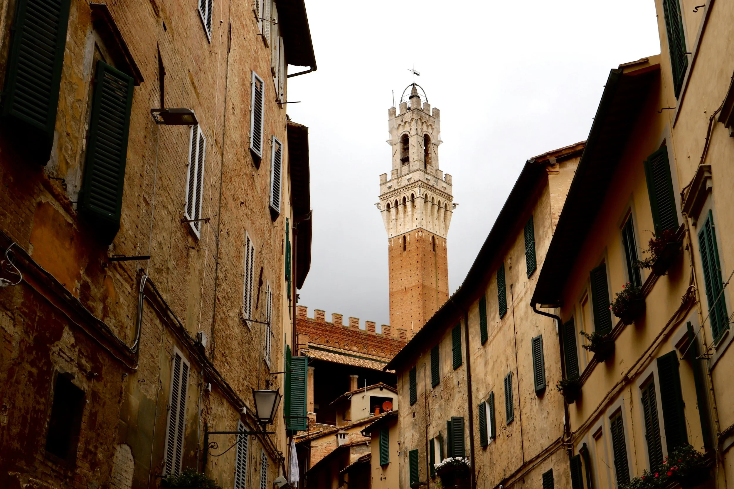 Narrow street with old brick buildings and green shutters, with a tall brick tower in the background.