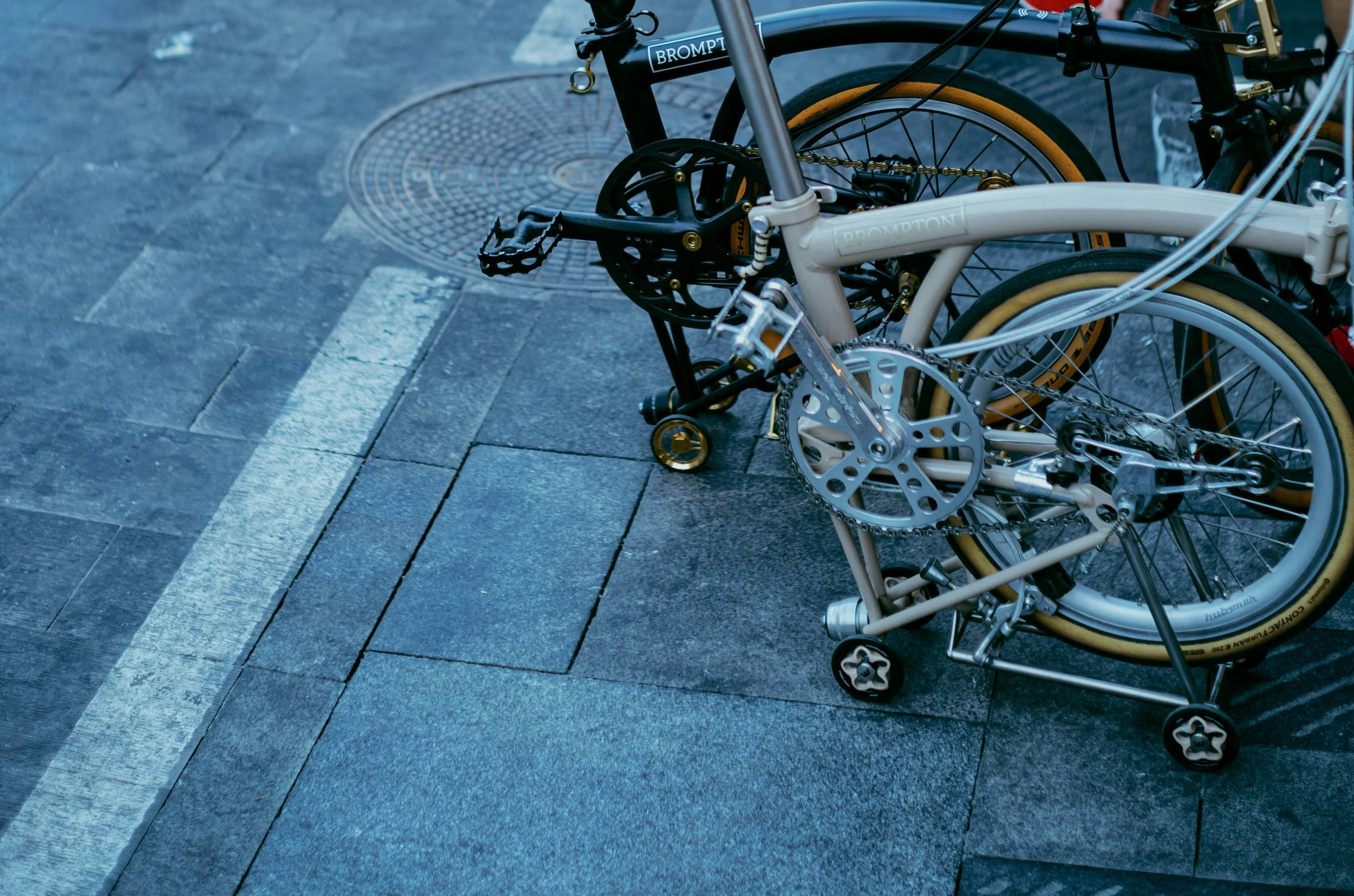 Brompton folding bicycle parked on a sidewalk with blue-gray paving stones and a white line painted on the ground.