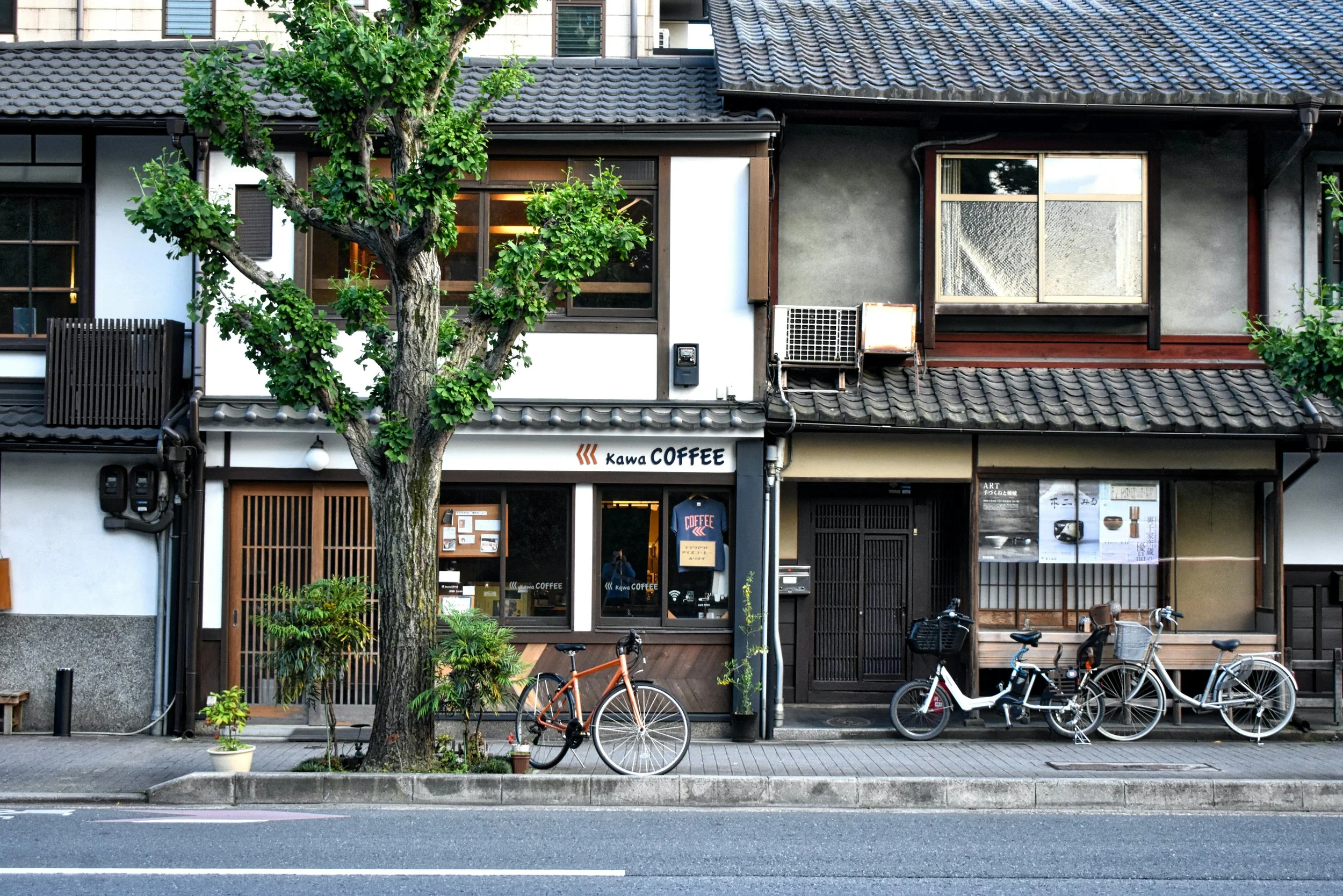 A street view of a cafe named Kawa Coffee with bicycles parked outside, a tree in front, and traditional Japanese-style buildings.