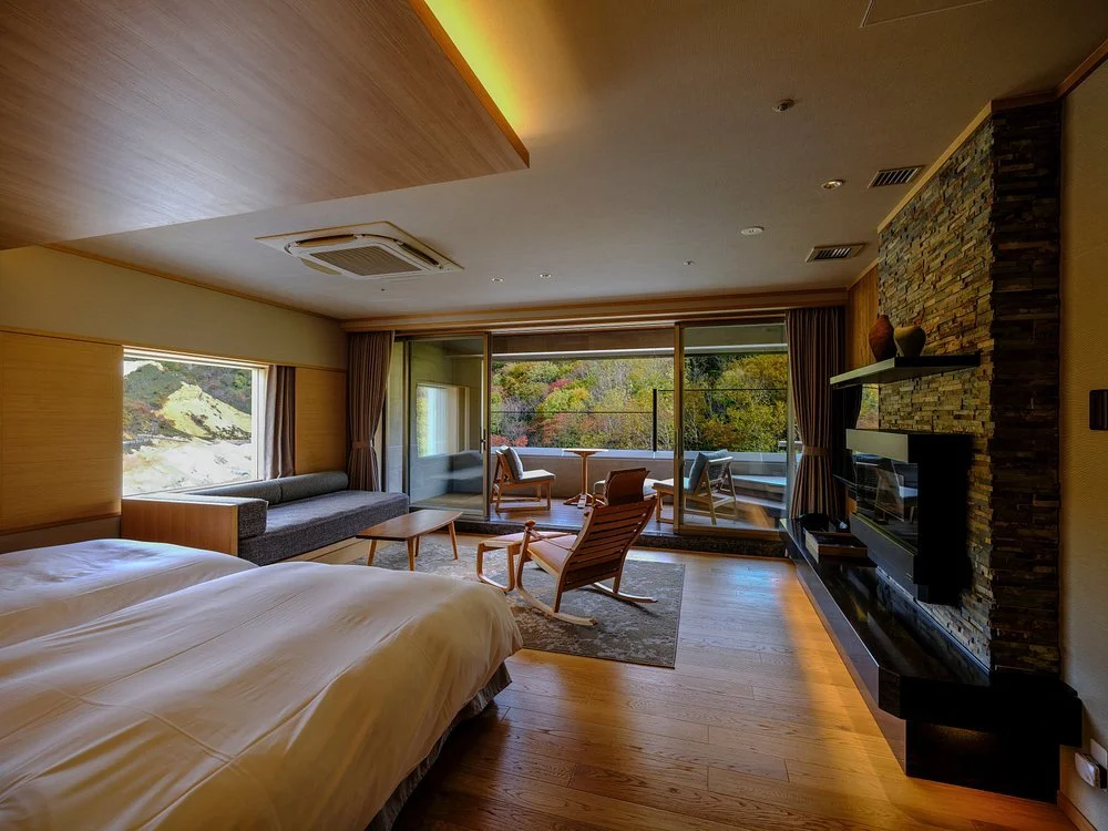 A modern hotel room with two beds, a sitting area by the window, and a balcony with outdoor furniture overlooking trees and hills.