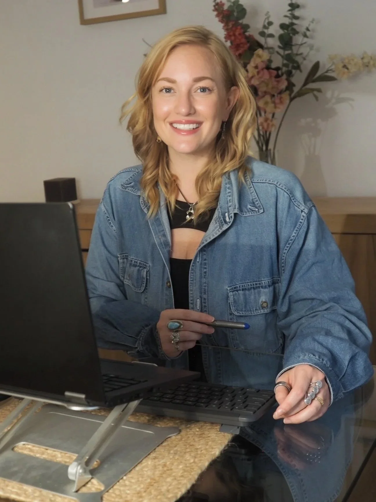 A woman with wavy blonde hair smiling while sitting at a desk with a laptop, wearing a denim jacket, rings, and earrings, with a vase of flowers in the background.