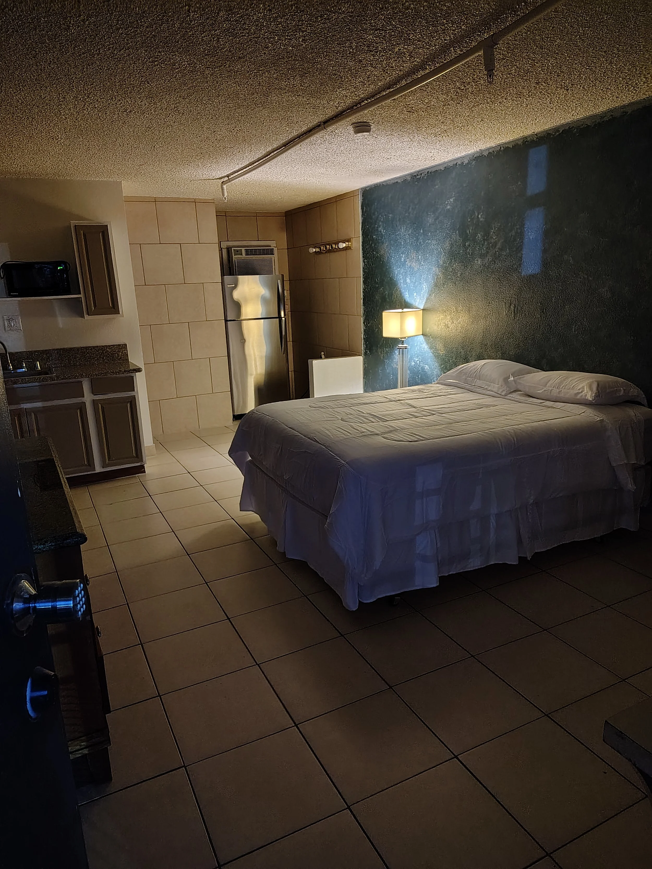 An interior hotel room with a bed, nightstand with lamp, kitchenette with refrigerator, microwave, cabinets, and ceramic tile flooring.