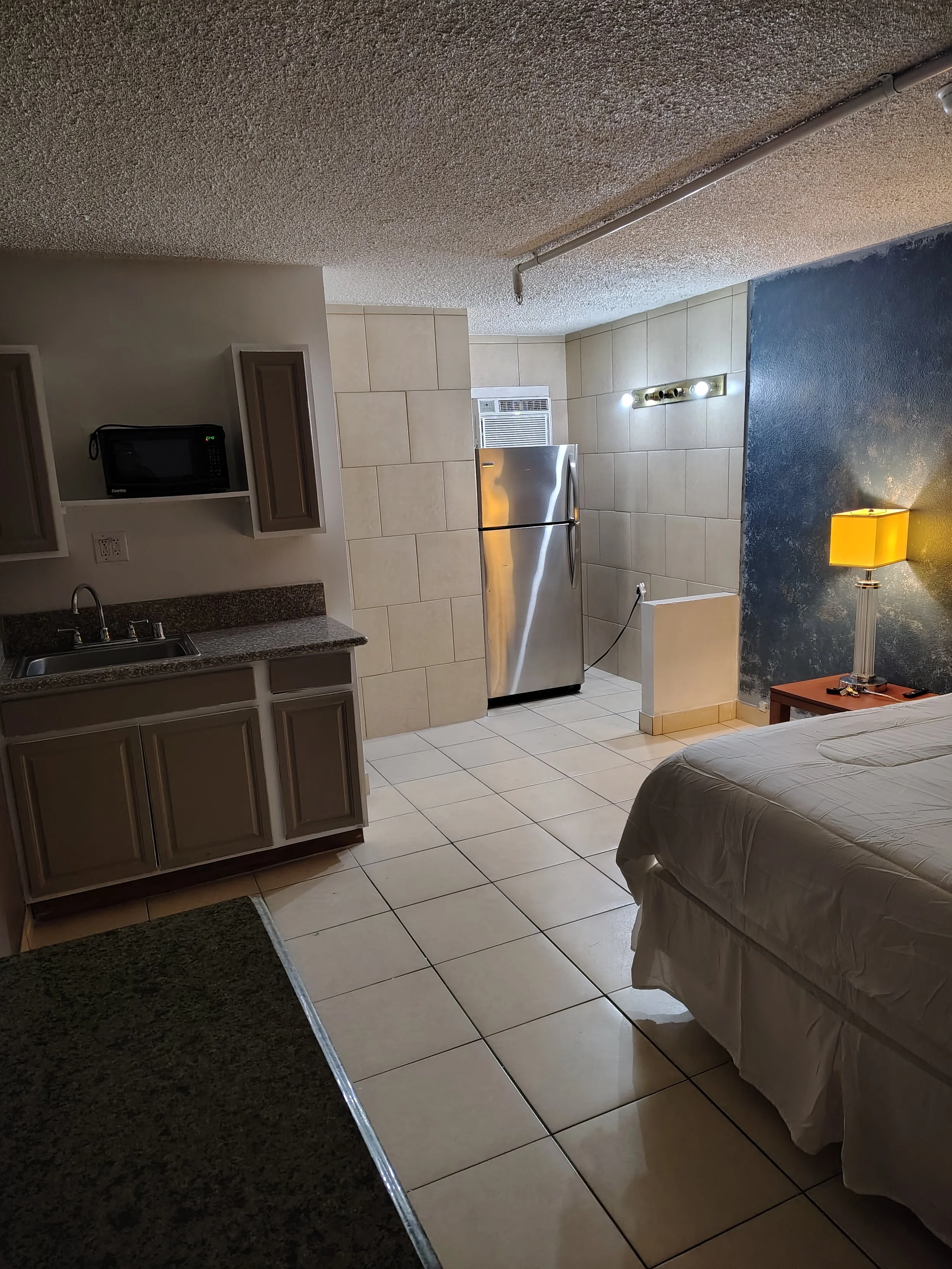 Interior of a modest hotel room or studio apartment featuring a bed partially visible on the right side, a nightstand with a yellow lamp, a kitchenette with cabinets, a microwave, a sink, and a small refrigerator, with tiled flooring and a textured c