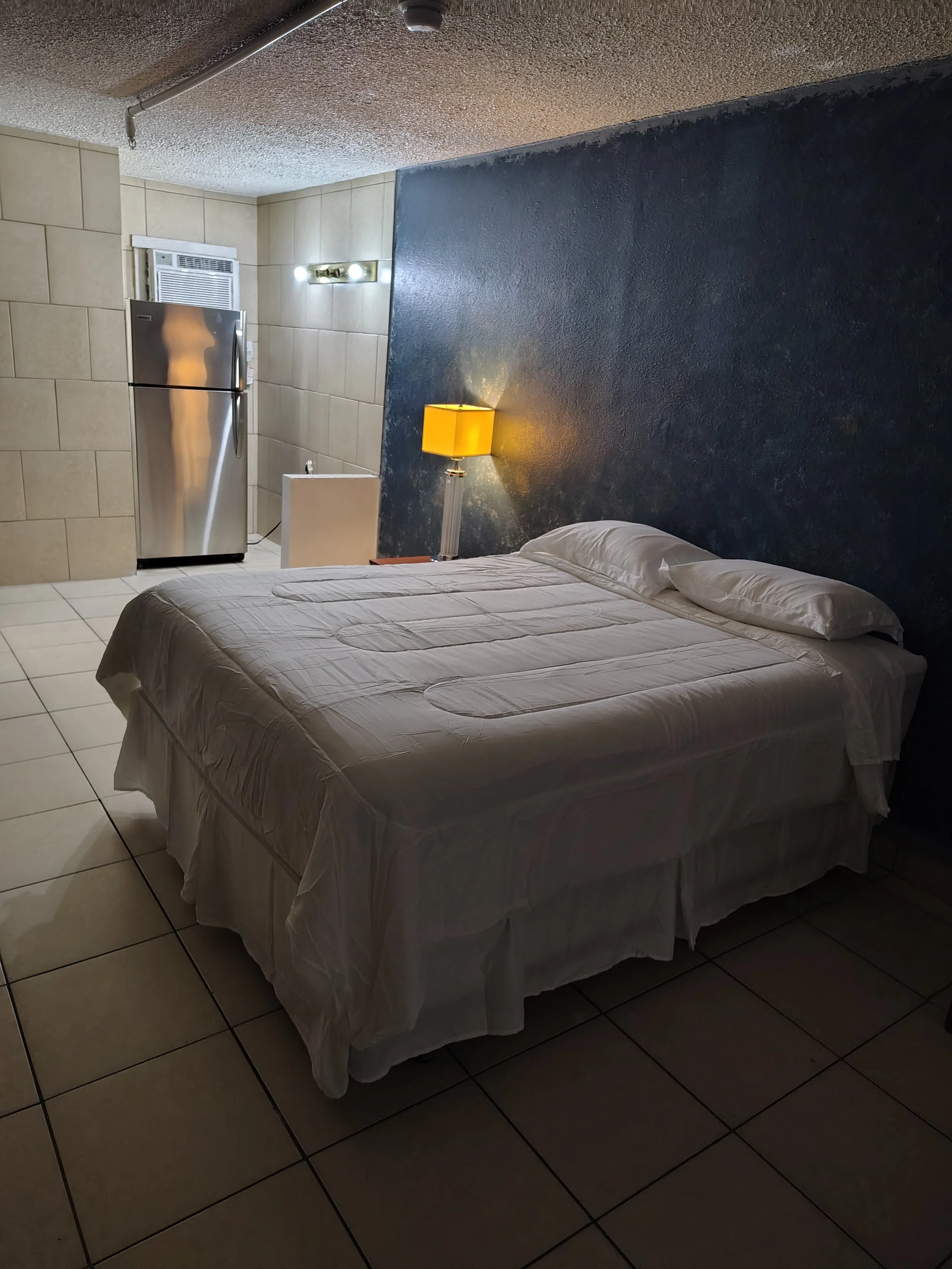 A bedroom with a white bed, pillows, a yellow bedside lamp, a small white table, and a mirrored refrigerator in the corner. The wall behind the bed is painted dark blue, and the room has tiled flooring.