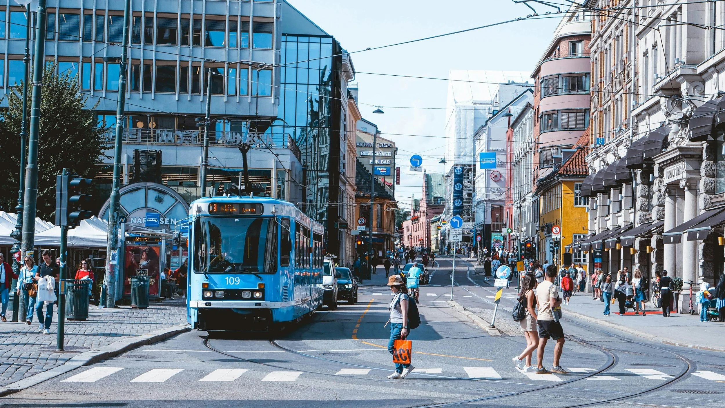 A street in Oslo, picturing a blu tram in the street, and a pedestrian with an orange bag crossing the street