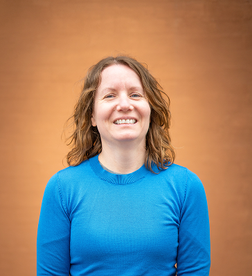 Brown haired woman in a bright blue shirt smiling at the camera in fron of an orange background; Marie Marqvardsen