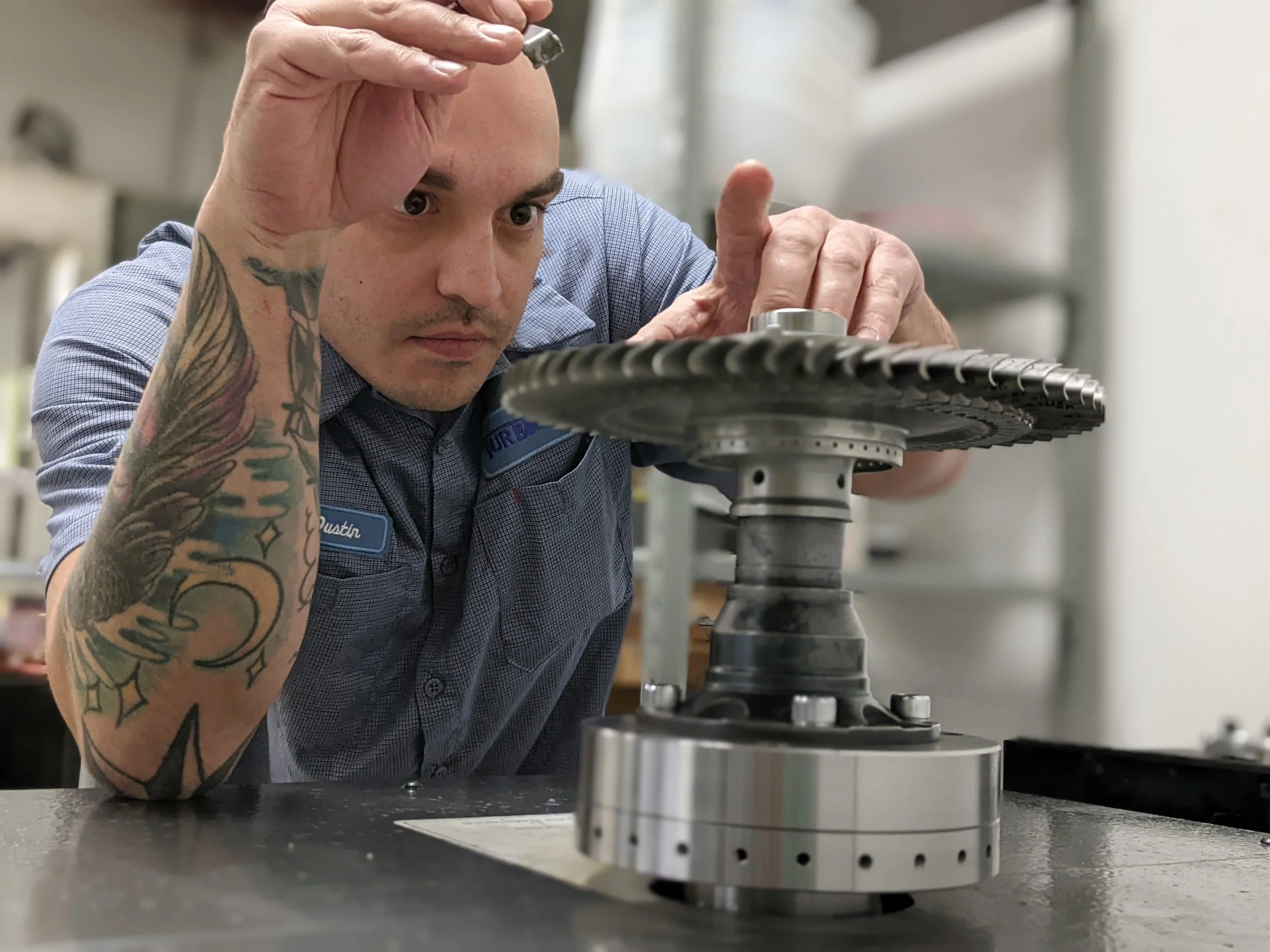 A man with tattoos on his arm, wearing a blue checkered shirt, is inspecting machinery or equipment on a workbench.