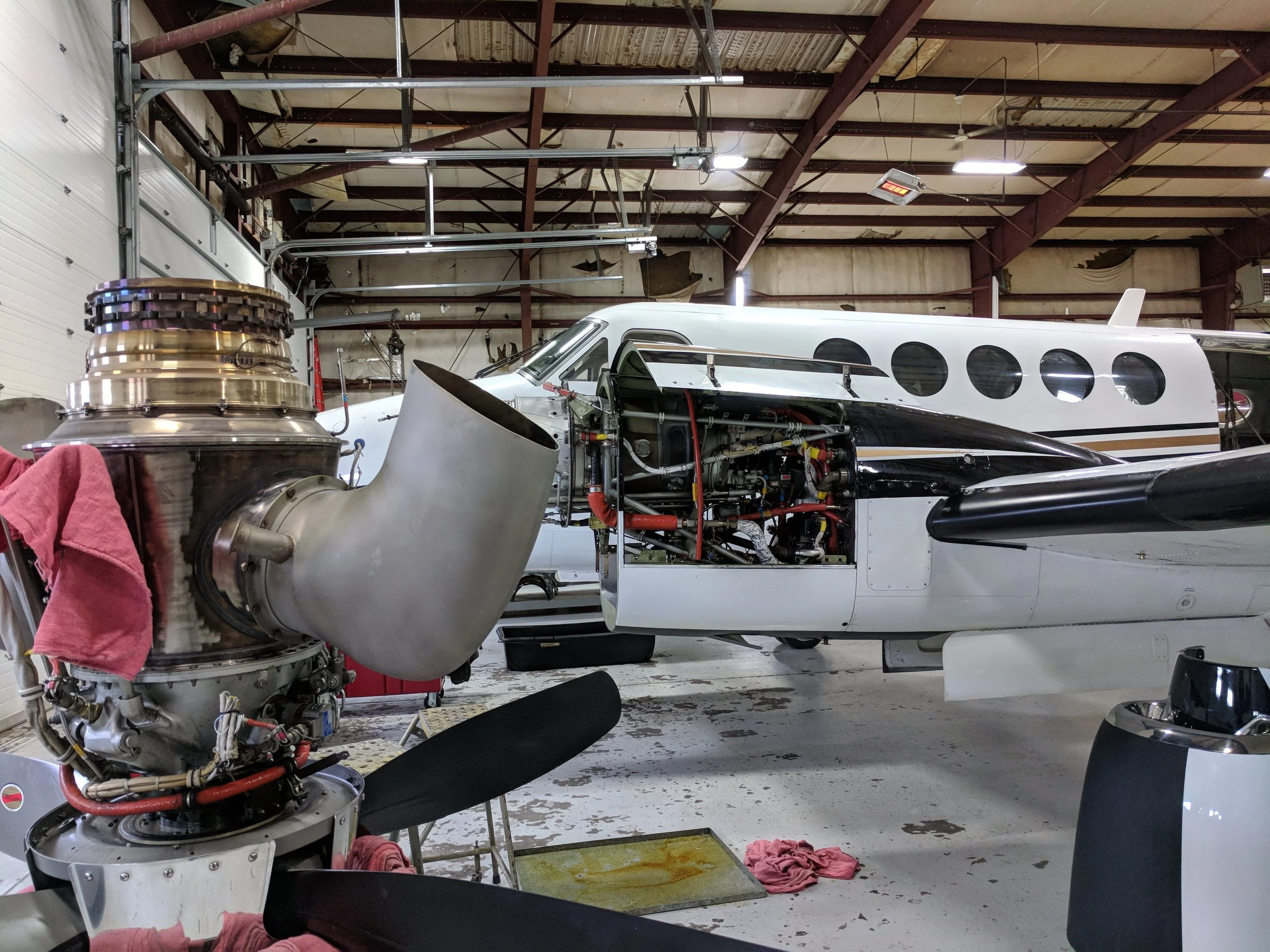 An aircraft cockpit with the fuselage open, revealing internal components and wiring, inside a hangar with a metal roof and some tools and cloths on the ground.
