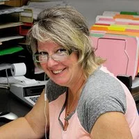 A smiling woman with glasses and gray hair sitting at a desk in an office, with colorful file folders and office supplies in the background.