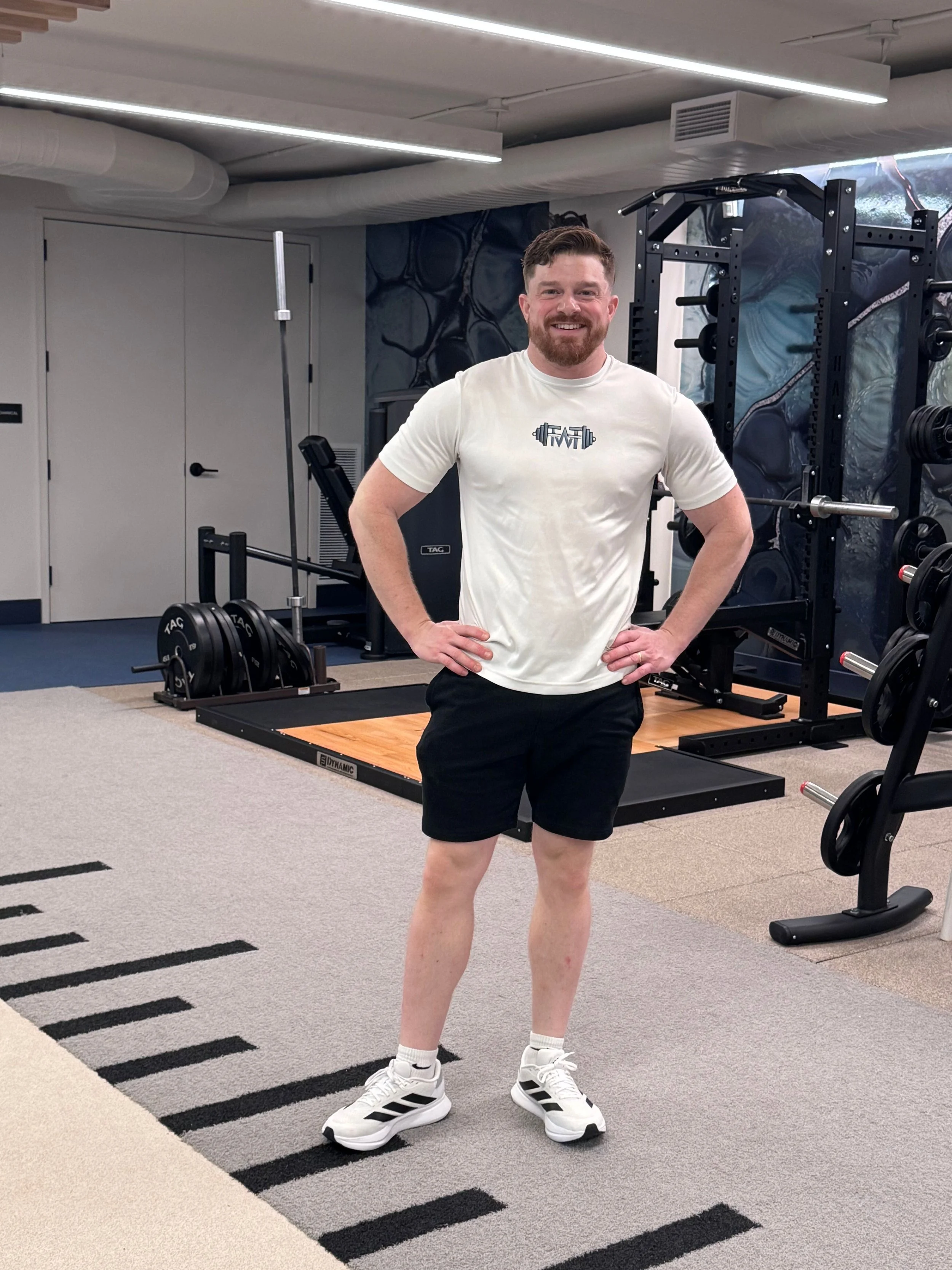 A personal trainer standing in a gym, smiling, wearing a white T-shirt with a barbell graphic, black shorts, and white sneakers with black stripes.