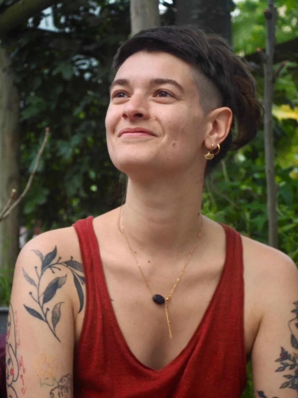 Une femme avec une coupe de cheveux courte et un côté rasé, portant un haut rouge, des bijoux en or et des tatouages sur les bras, souriant dans un environnement extérieur avec des plantes en arrière-plan.