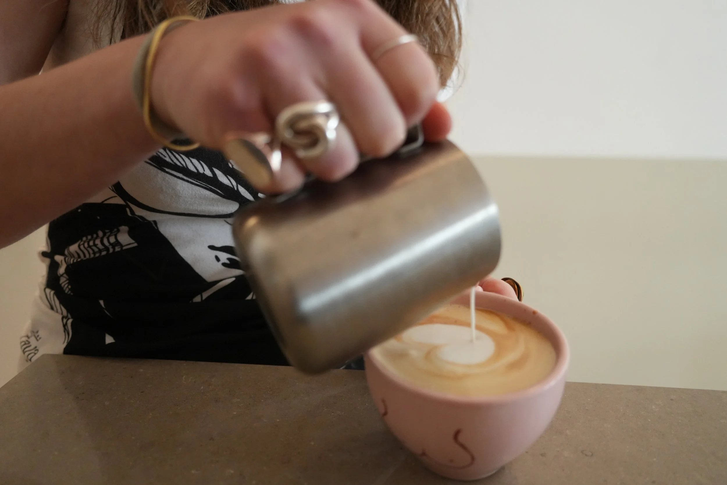 Une main avec des bagues verse un liquide dans une tasse rose, vue de côté.