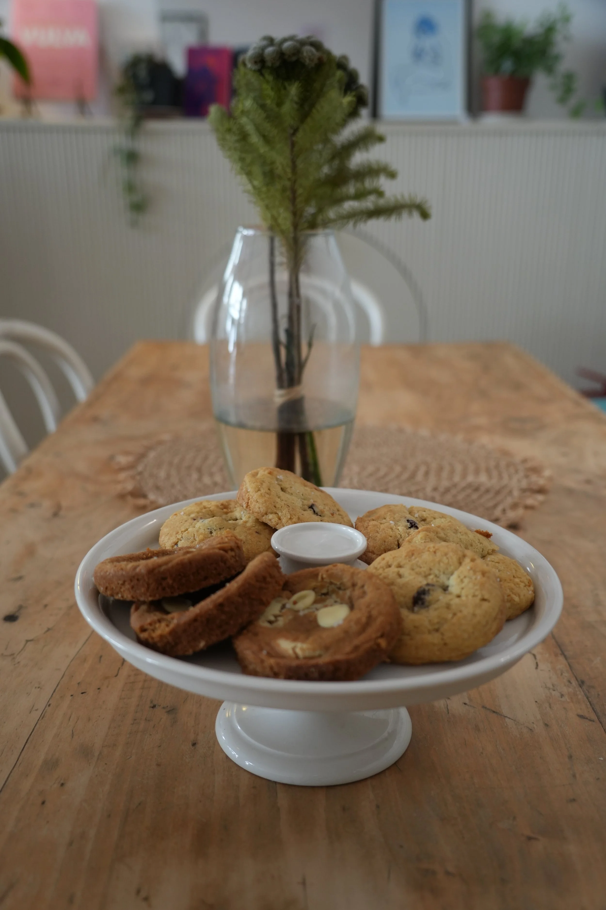 Plateau de biscuits variés sur une table en bois, avec une assiette de cookies et autres biscuits, et un vase avec une plante verte en arrière-plan.