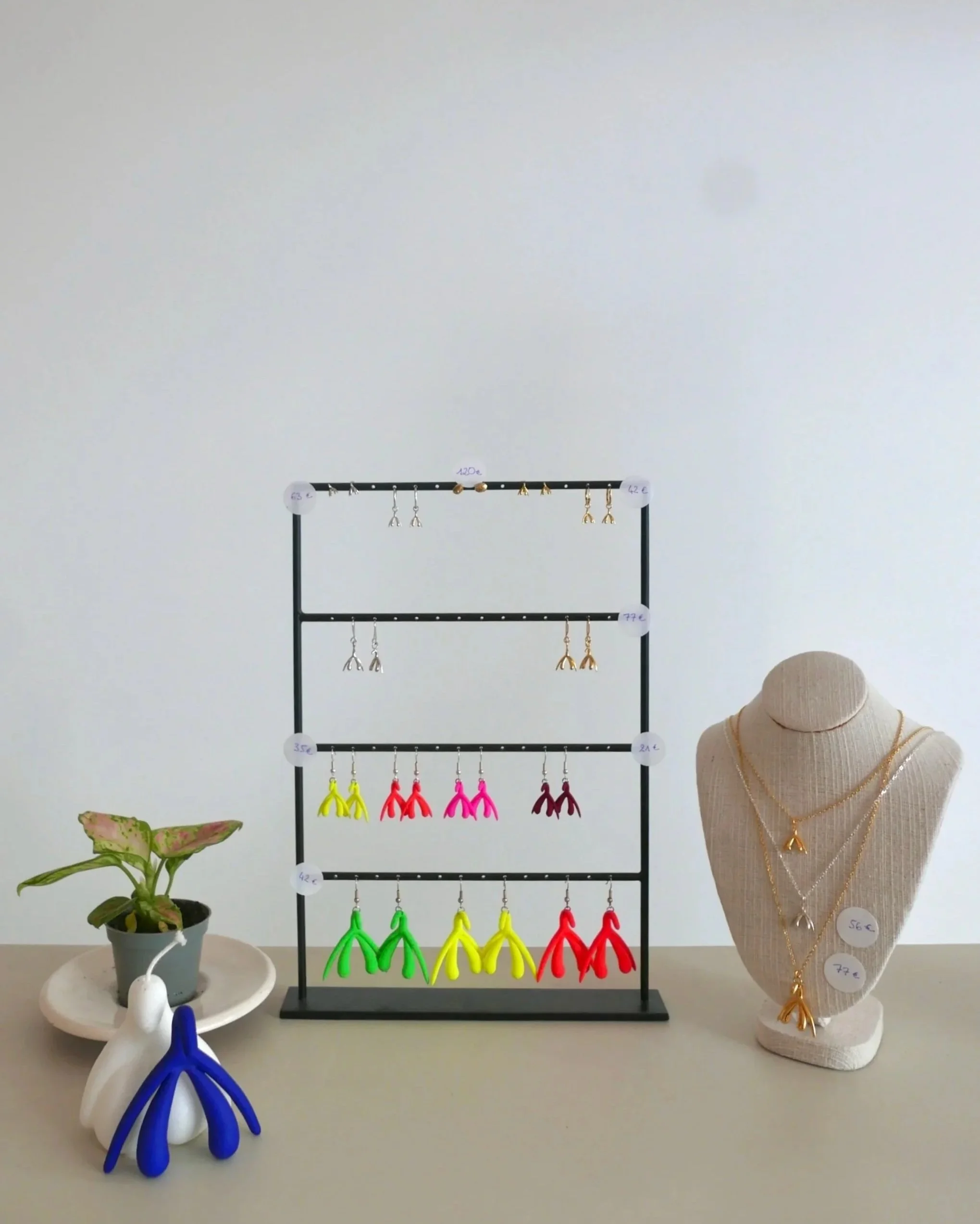 Un présentoir à bijoux avec des boucles d'oreilles colorées, une statue en forme de collier avec des colliers dorés, une plante verte en pot, et des objets en forme de mains ou de tortues en plastique. Le fond est un mur blanc, et il y a une chaise en partie visible sur le côté droit.