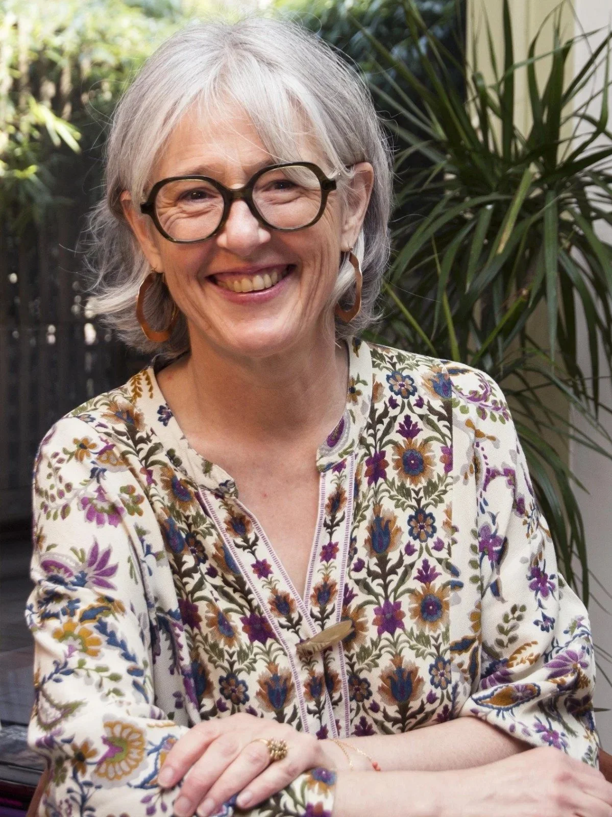 Une femme âgée souriante avec des lunettes rondes et des boucles d'oreilles en forme de cercle, portant une chemise florale colorée, assise à une table avec un fond de plantes vertes dans une atmosphère claire et naturelle.