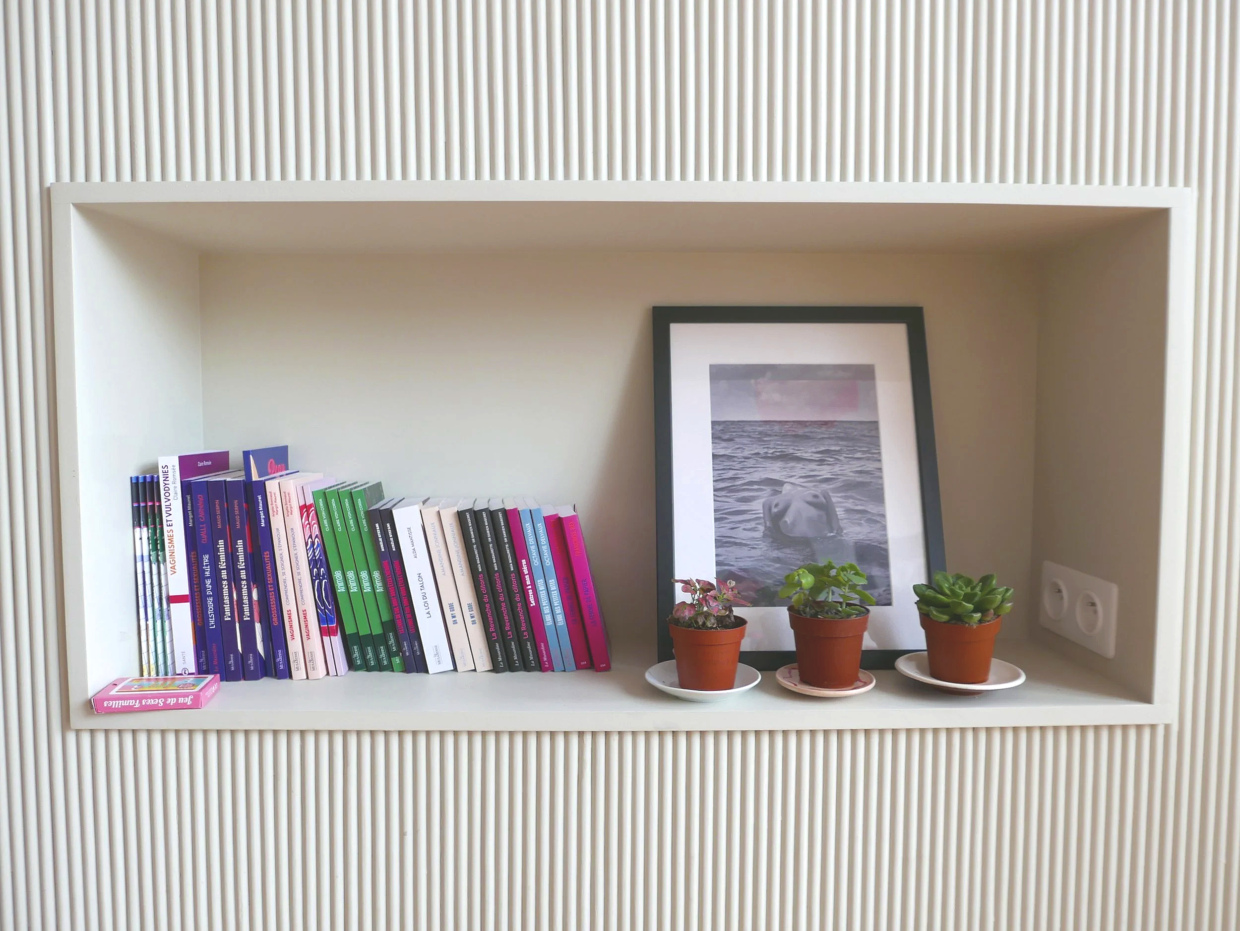 Une étagère blanche avec des livres colorés, un cadre avec une photo de la mer, deux petites plantes en pot et un plateau blanc. La façade de l'étagère a un motif rayé vertical.