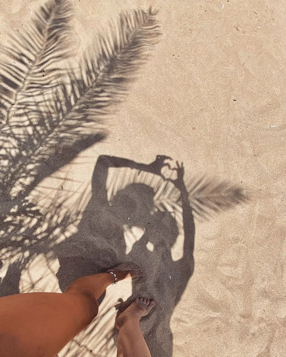 Shadow of a person taking a photo, with one foot on the sand and palm tree leaves casting a shadow on the ground.