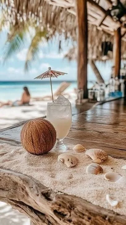 A tropical beach scene with a drink topped with ice and a small umbrella, a coconut, and seashells on a wooden surface, with the ocean and a person relaxing on the sand in the background.