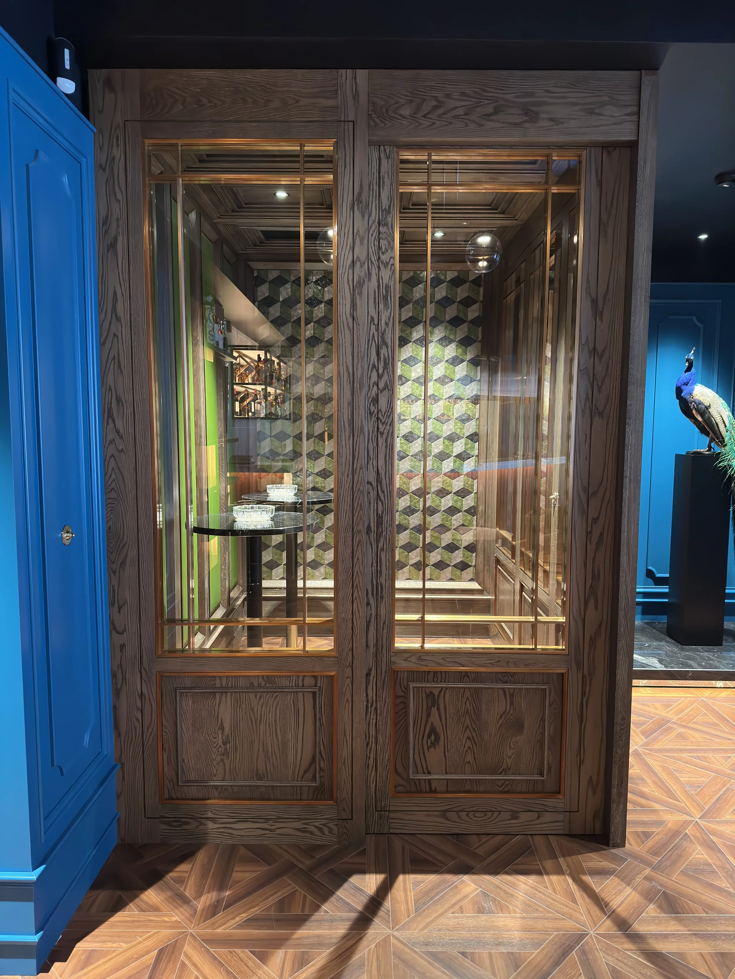 A wooden framed glass display cabinet with gold accents, showcasing a patterned wallpaper background and a black table inside. To the left, a blue cabinet is partially visible.