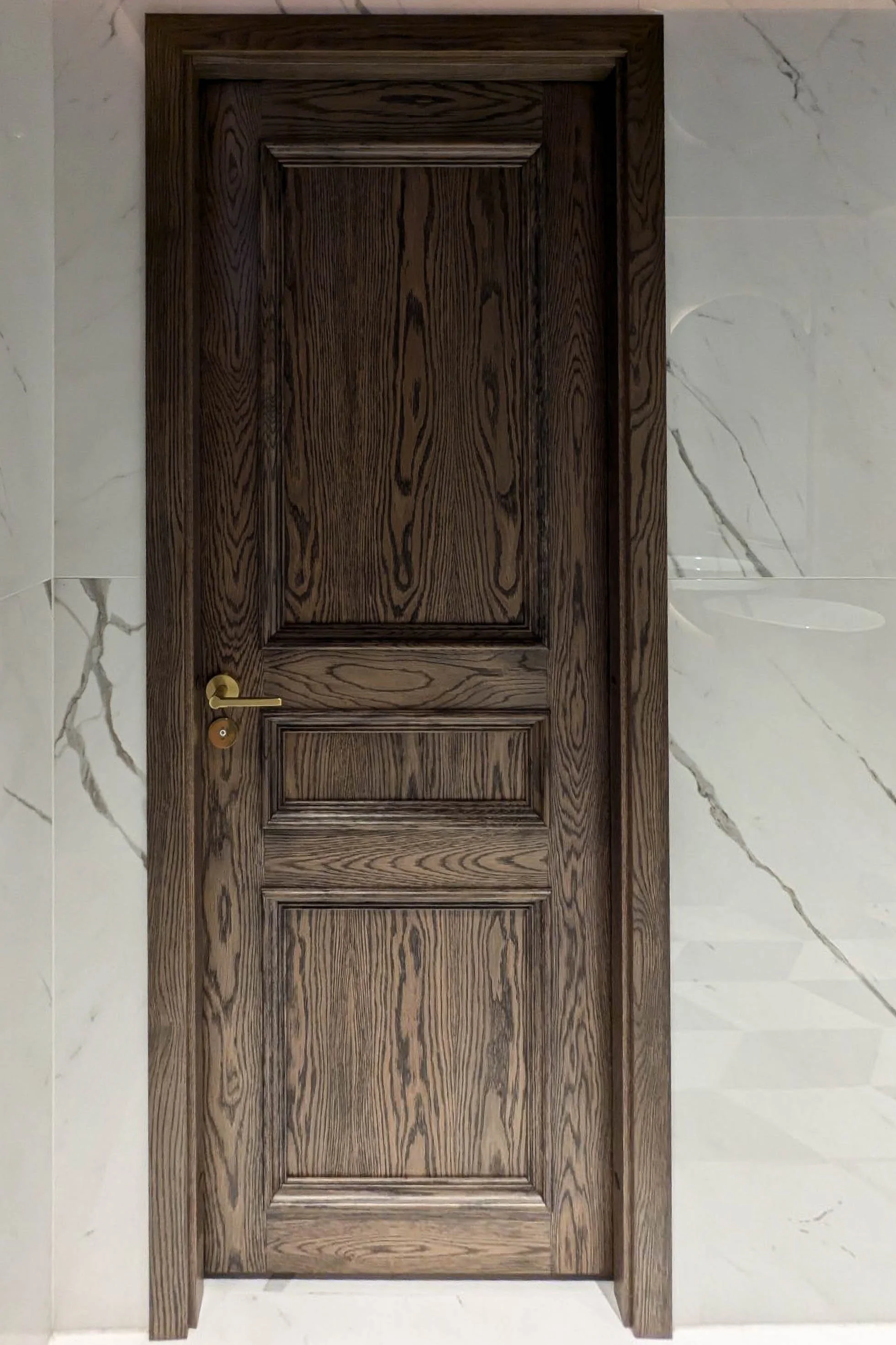A wooden door with a gold handle, set in a wall with white marble tiles with gray veining.