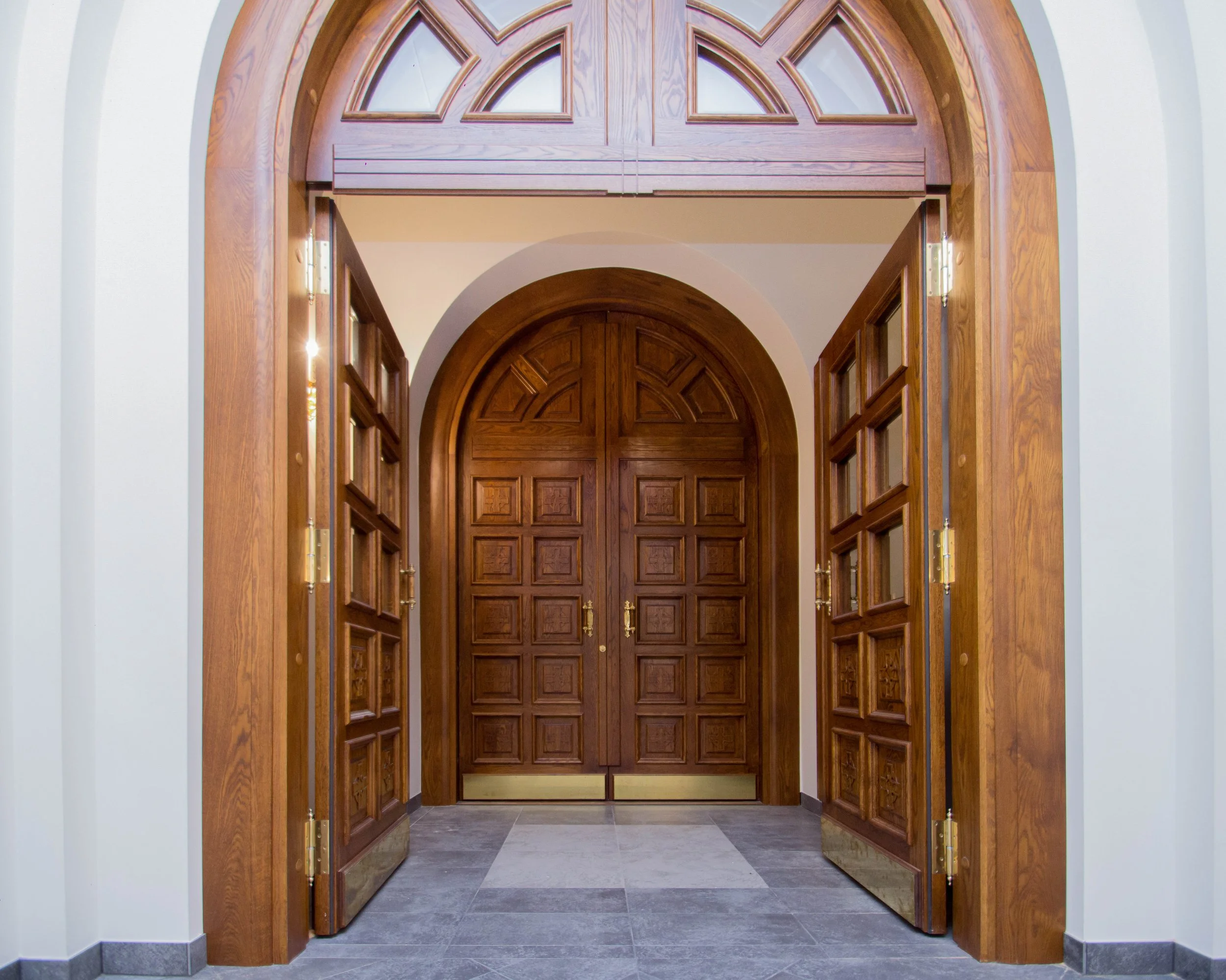 Wooden double doors with brass handles, surrounded by a wooden frame with decorative glass panels above, leading into a building with a tile floor.
