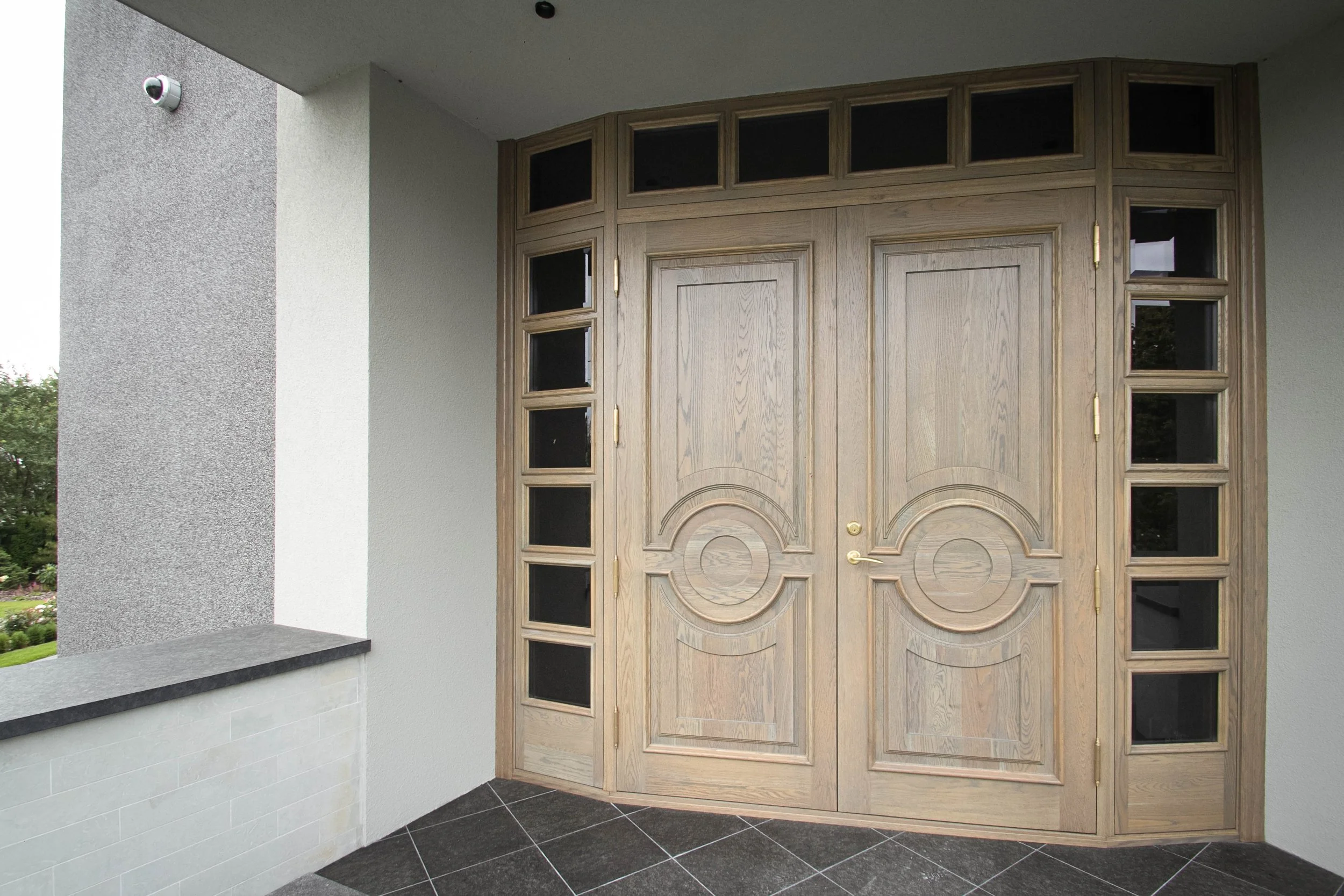 Double wooden front doors with decorative patterns, surrounded by multiple small window panels, on an outdoor porch with gray tiled flooring and textured gray and white walls.