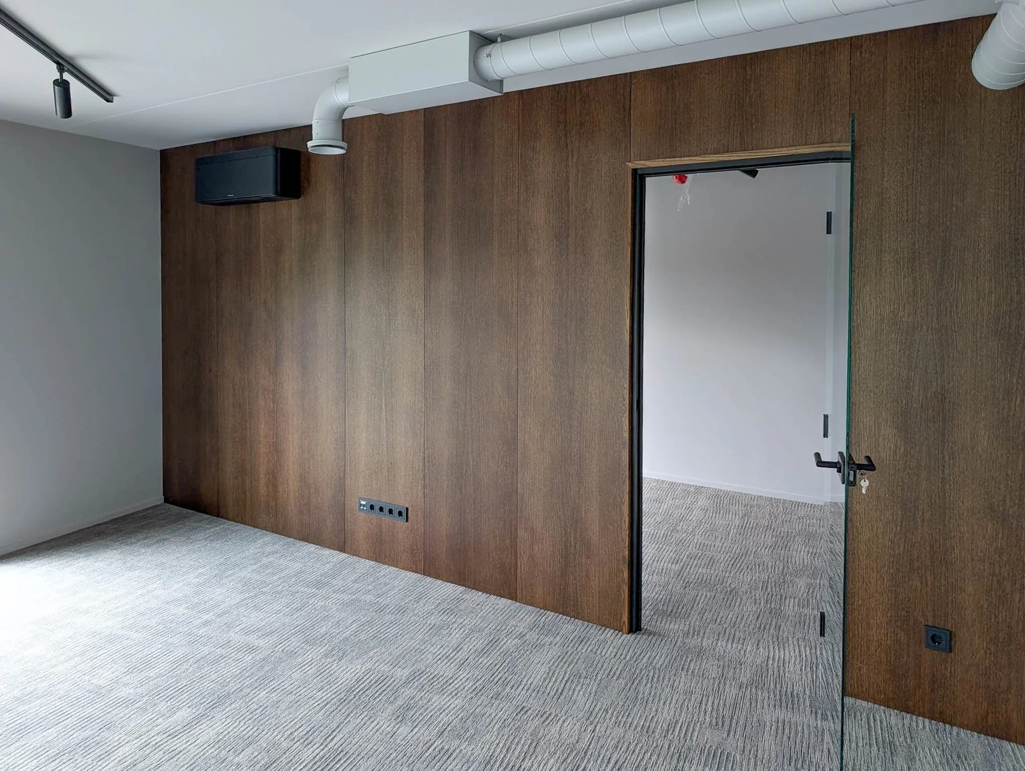 Empty room with wood-paneled wall, carpeted floor, glass door, black speaker mounted near ceiling, ceiling-mounted spotlight, and several electrical outlets.