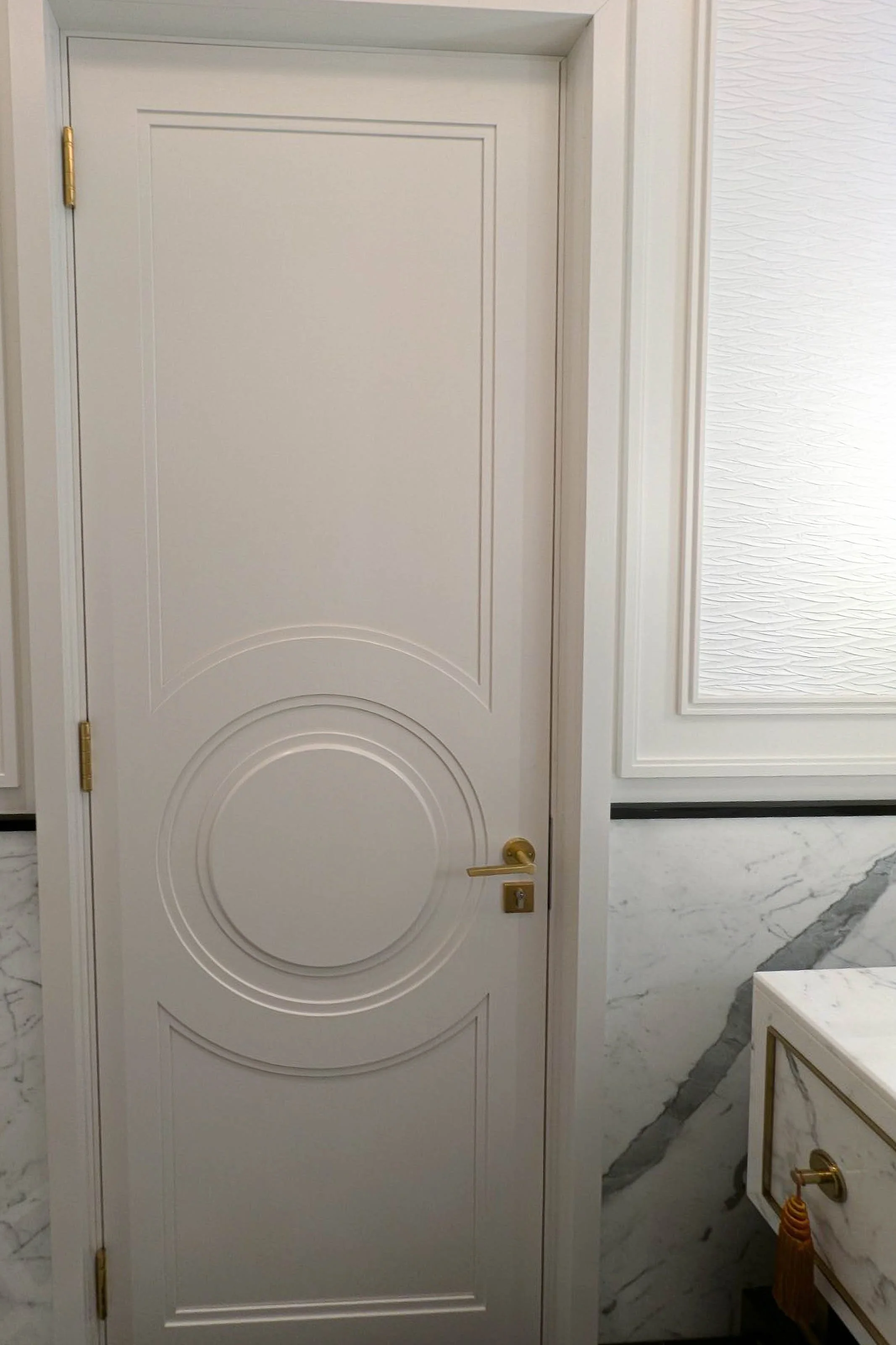 A white door with decorative circular and rectangular carvings, gold hinges, and a gold door handle, set in an elegant interior with marble walls and trim.