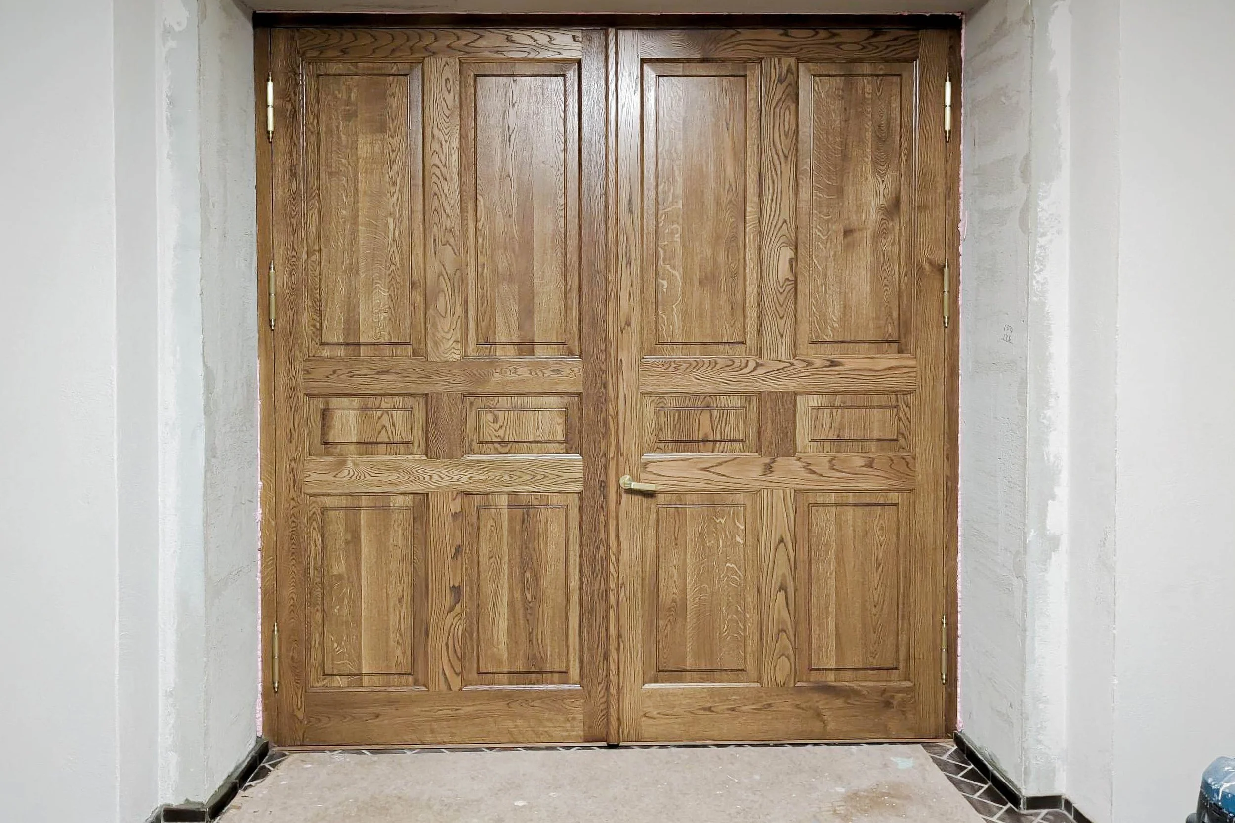 Wooden double door with gold hinges and handle, partially surrounded by unfinished walls and a tiled floor.