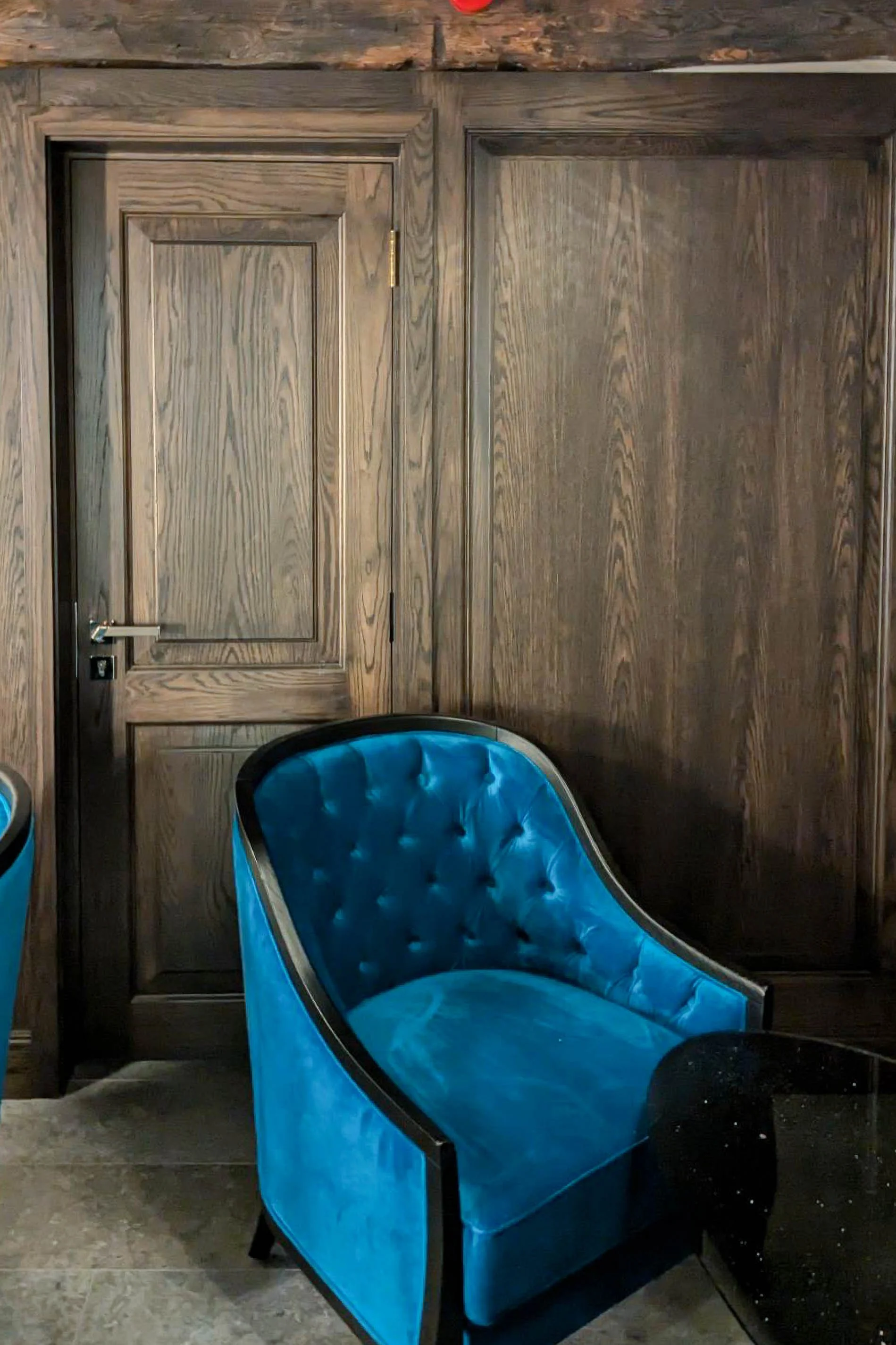 A bright blue velvet armchair with a black frame is positioned against a wooden wall with a matching door in the background.