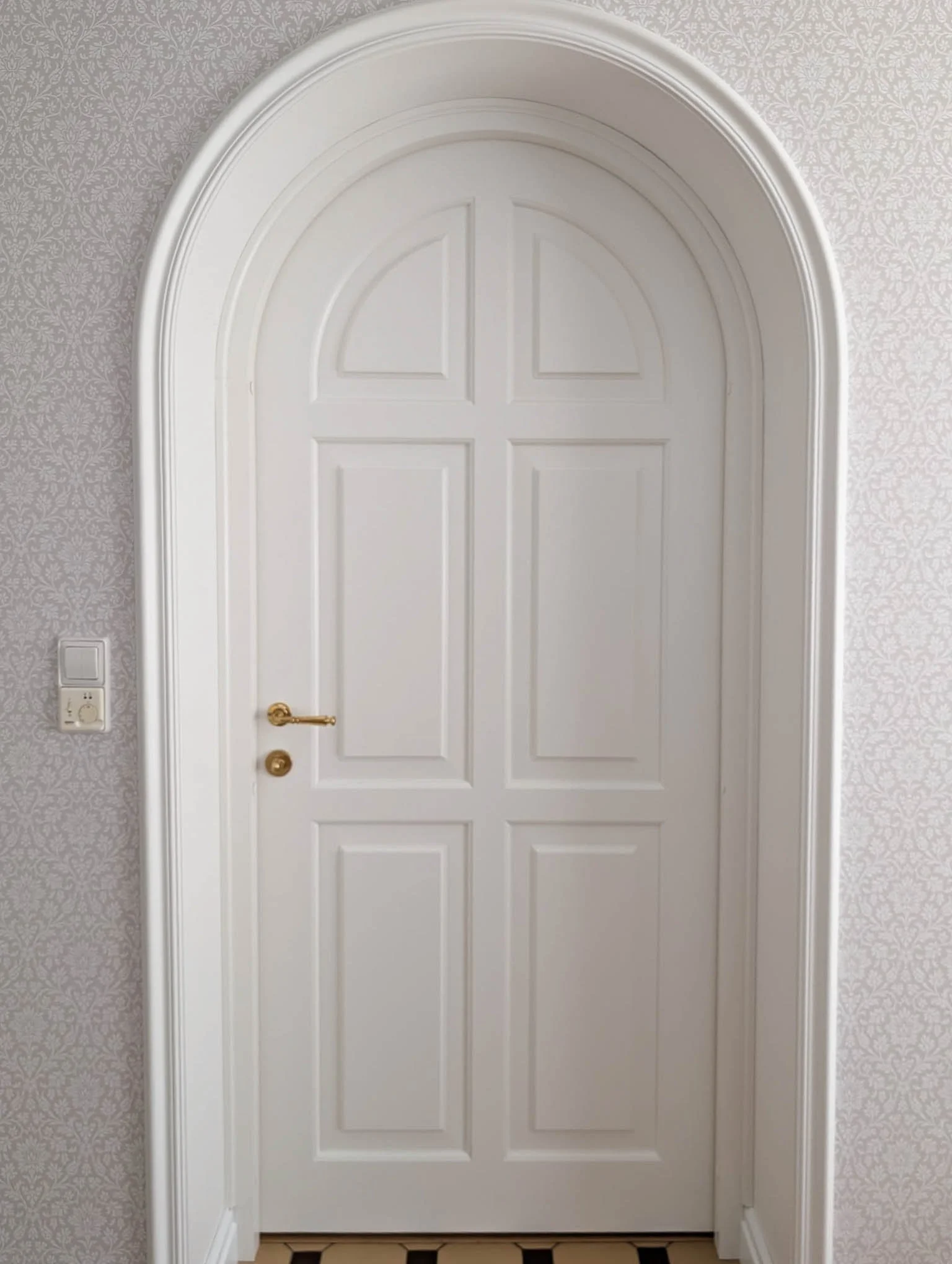 White arched interior door with panel detailing, gold handle, set in a wall with light patterned wallpaper.