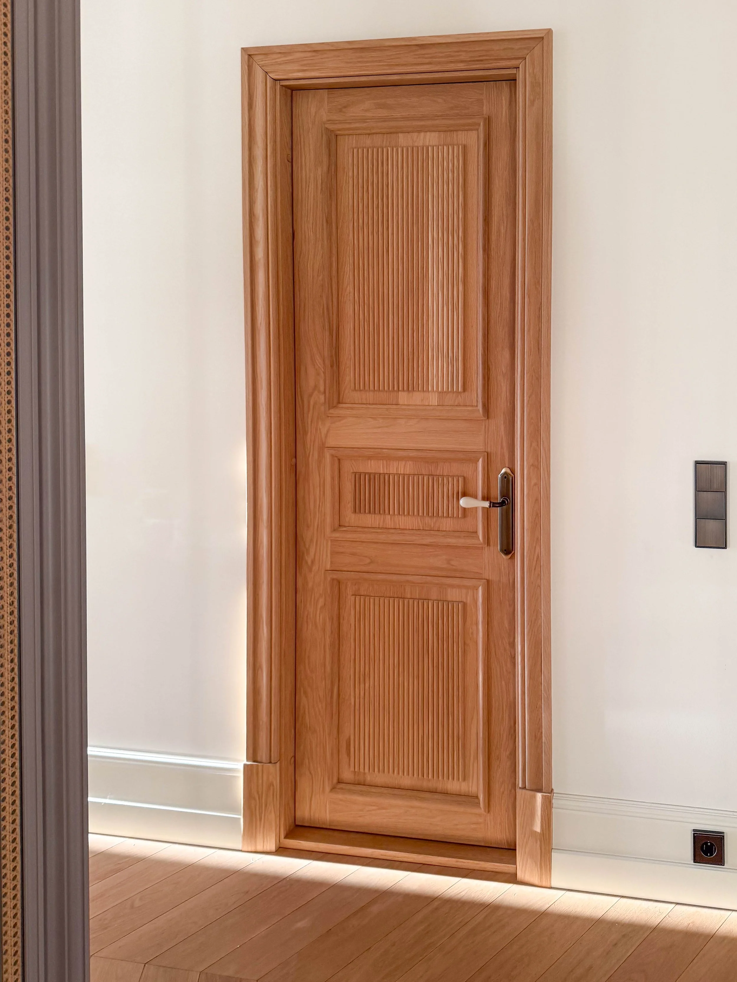 A wooden interior door with a brass handle and vertical beadboard panels, set in a white wall, with a light wood floor and a black power outlet nearby.