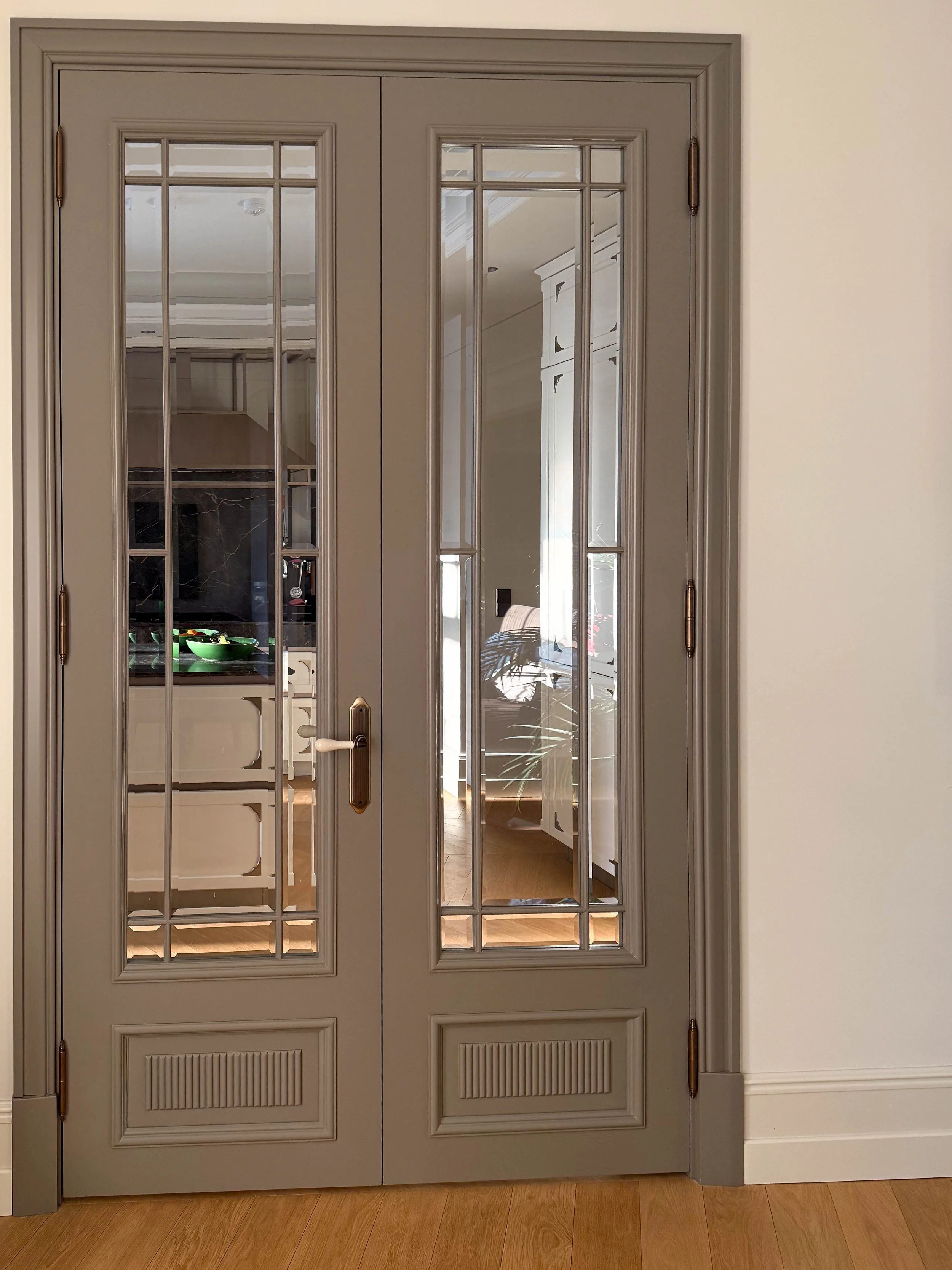 Double glass-paned French doors with beige frames and brass hardware, leading into a kitchen and living area.