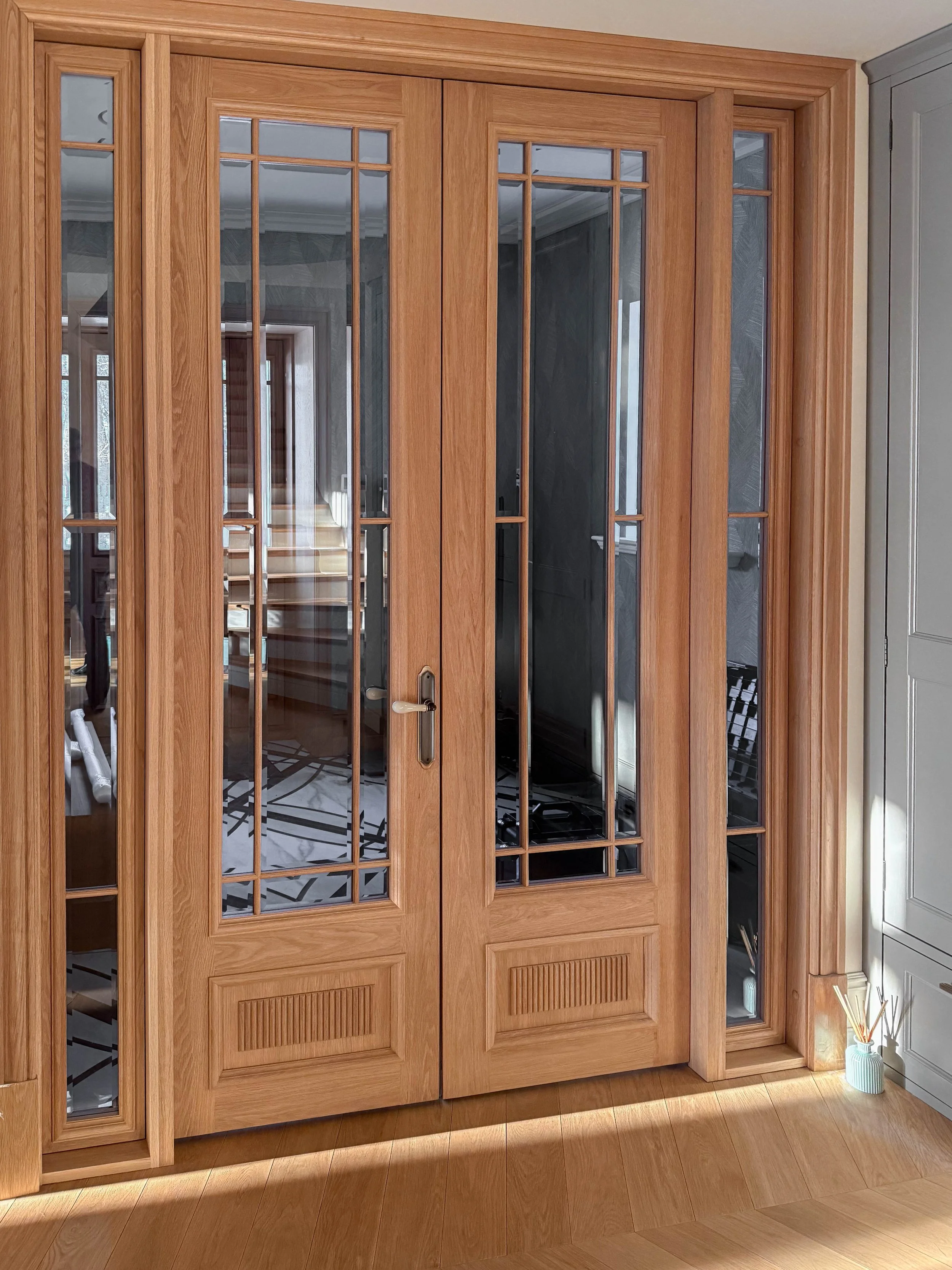 Wooden glass-paneled double doors with decorative wooden trim, reflected in the glass, leading into a room with staircase and patterned floor tiles. Light wood flooring and a small vase with reed diffusers are visible nearby.