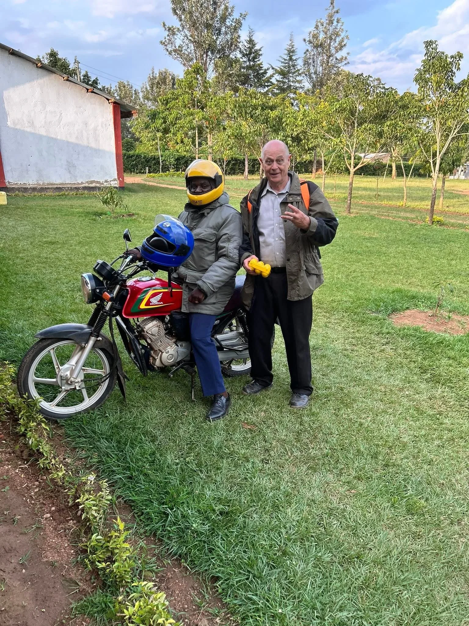 Two men standing outdoors on grass, one sitting on a red motorcycle wearing a helmet, the other making a peace sign, with trees and a white building in the background.