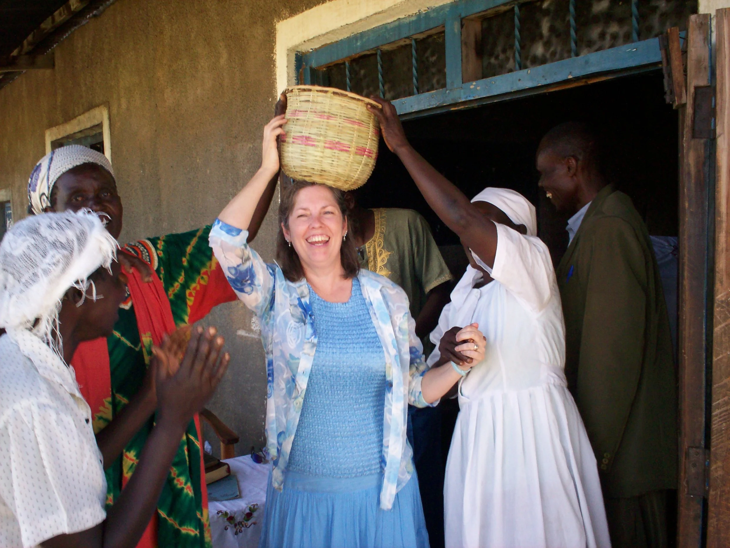 A group of women, including a woman in a blue dress, celebrate a special moment as she is crowned with a basket by another woman. The group is smiling and appears joyful, standing outside a building.