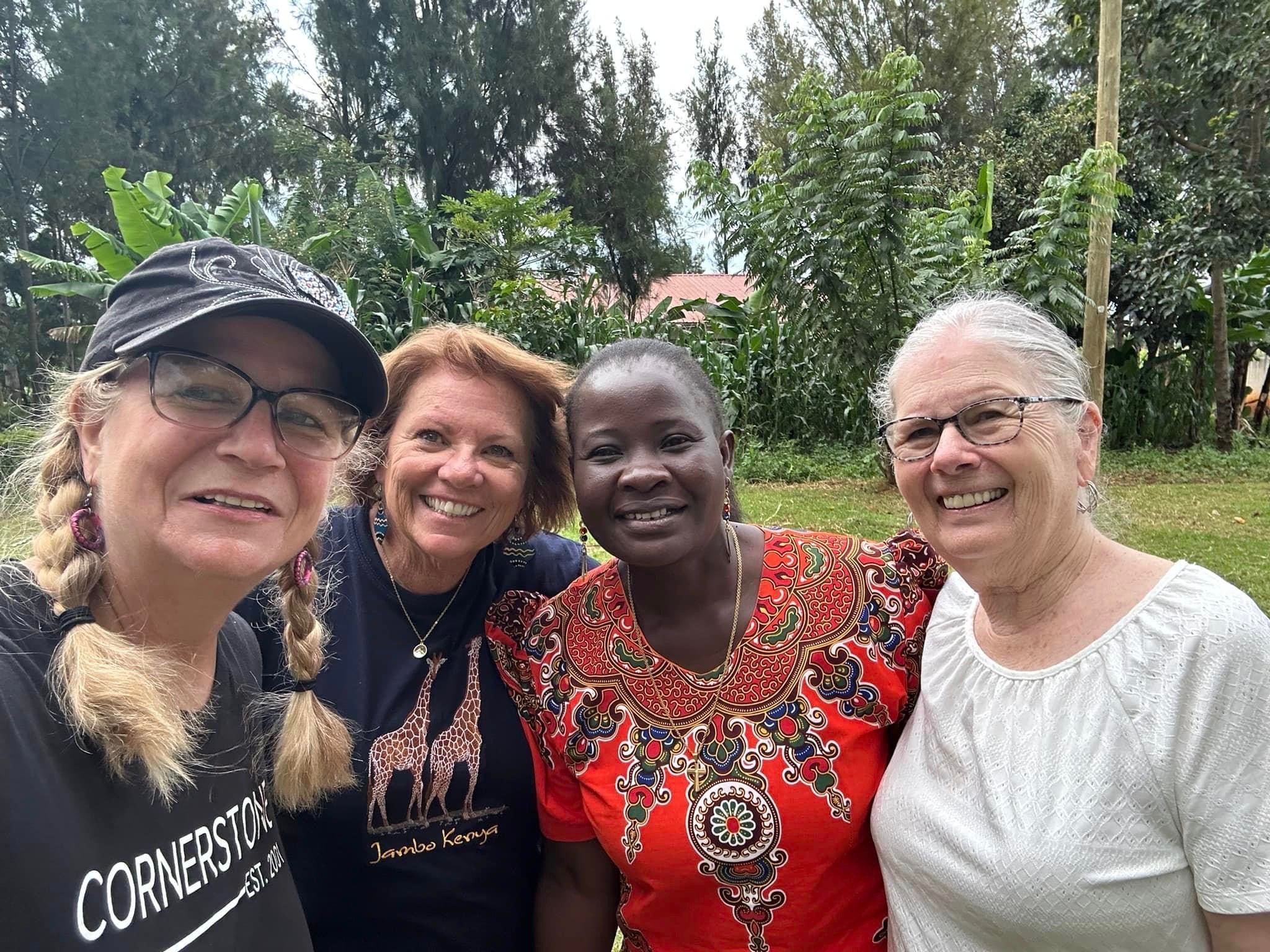 Four women smiling outdoors with trees and greenery in the background.
