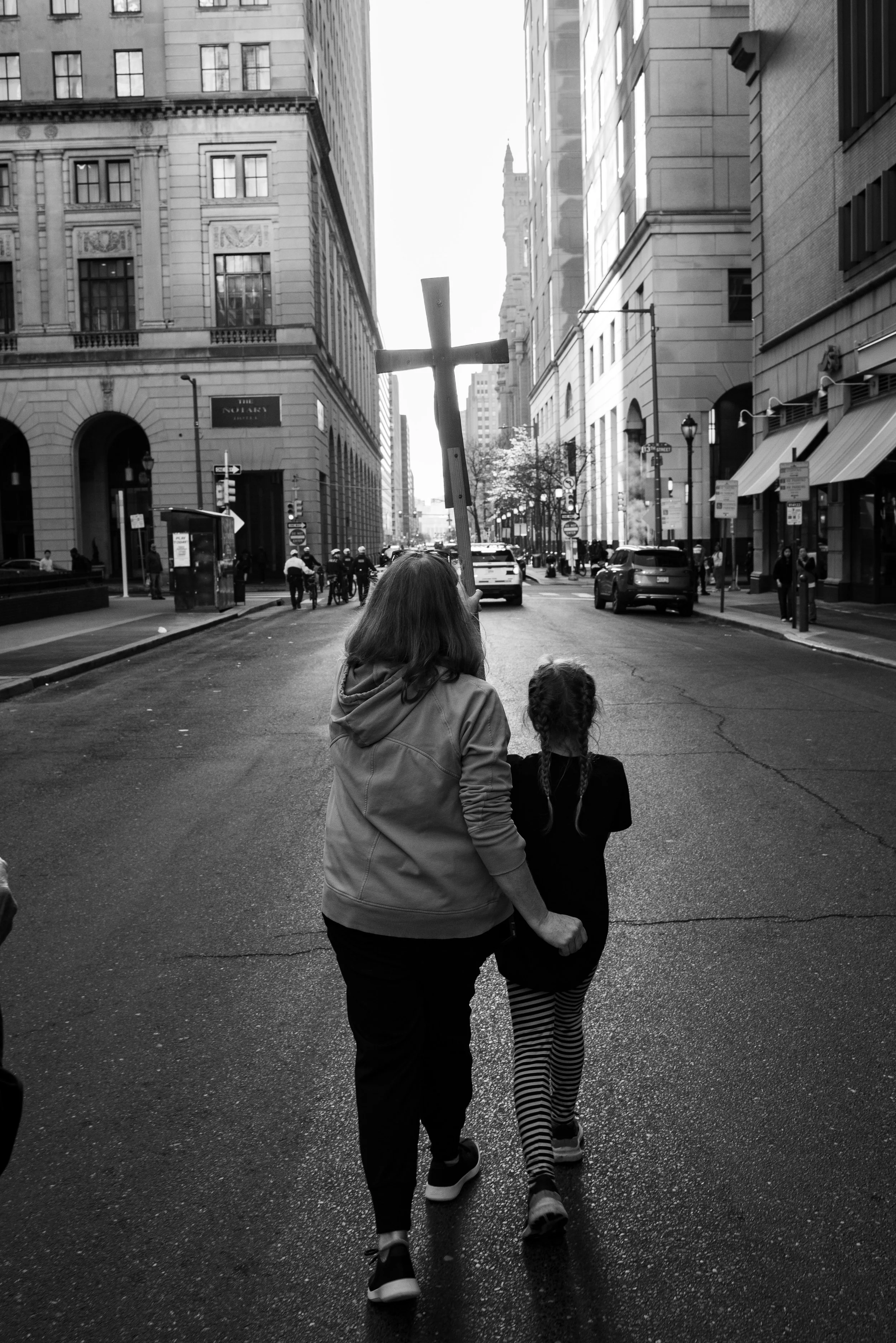 A woman and a young kid walking together down a city street, with the woman holding a large cross on a stick. The scene is in black and white, with tall buildings on both sides and several people and cars visible on the street.