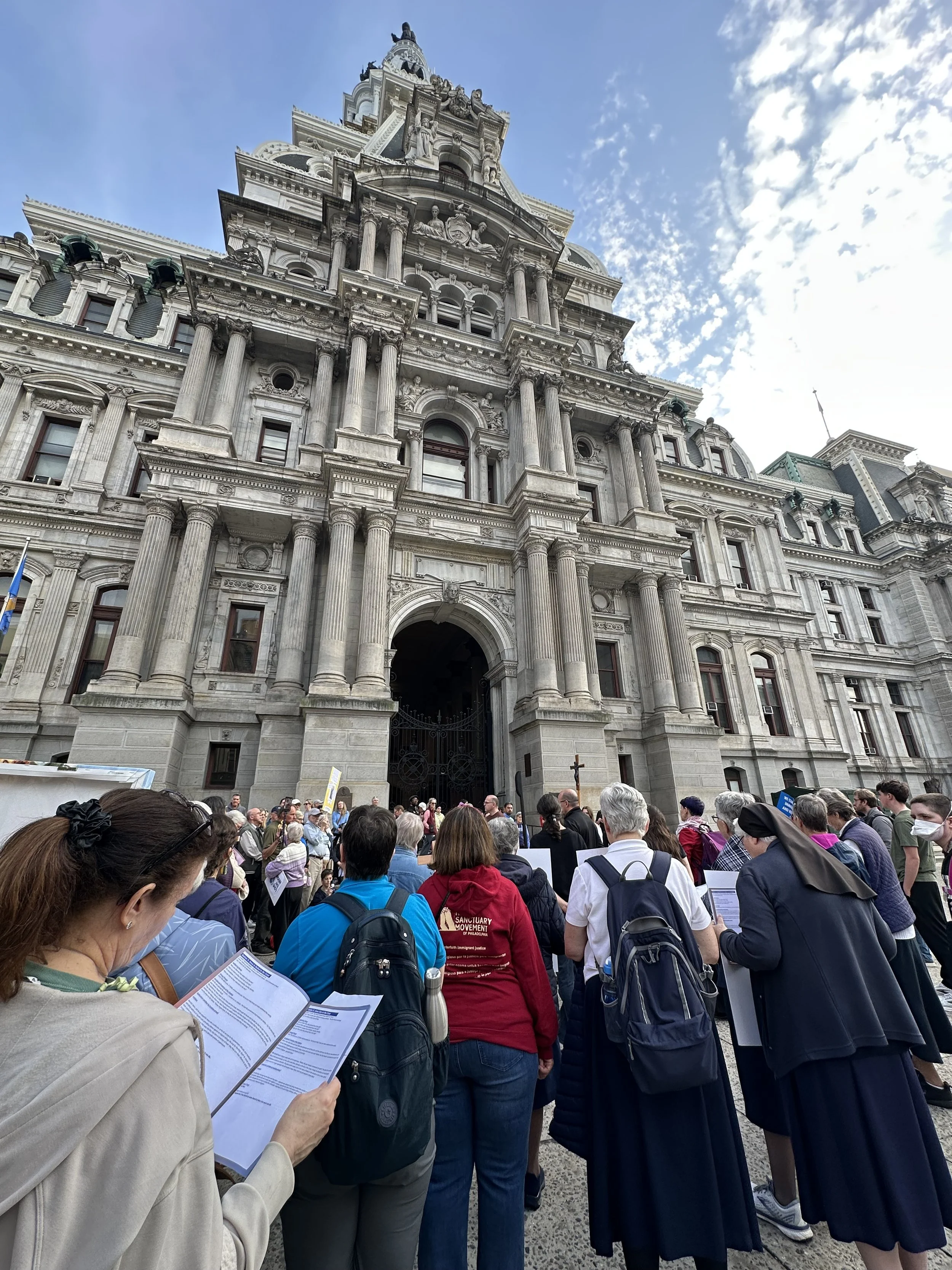 Crowd gathered outside Philadelphia City Hall during daytime with a partly cloudy sky.