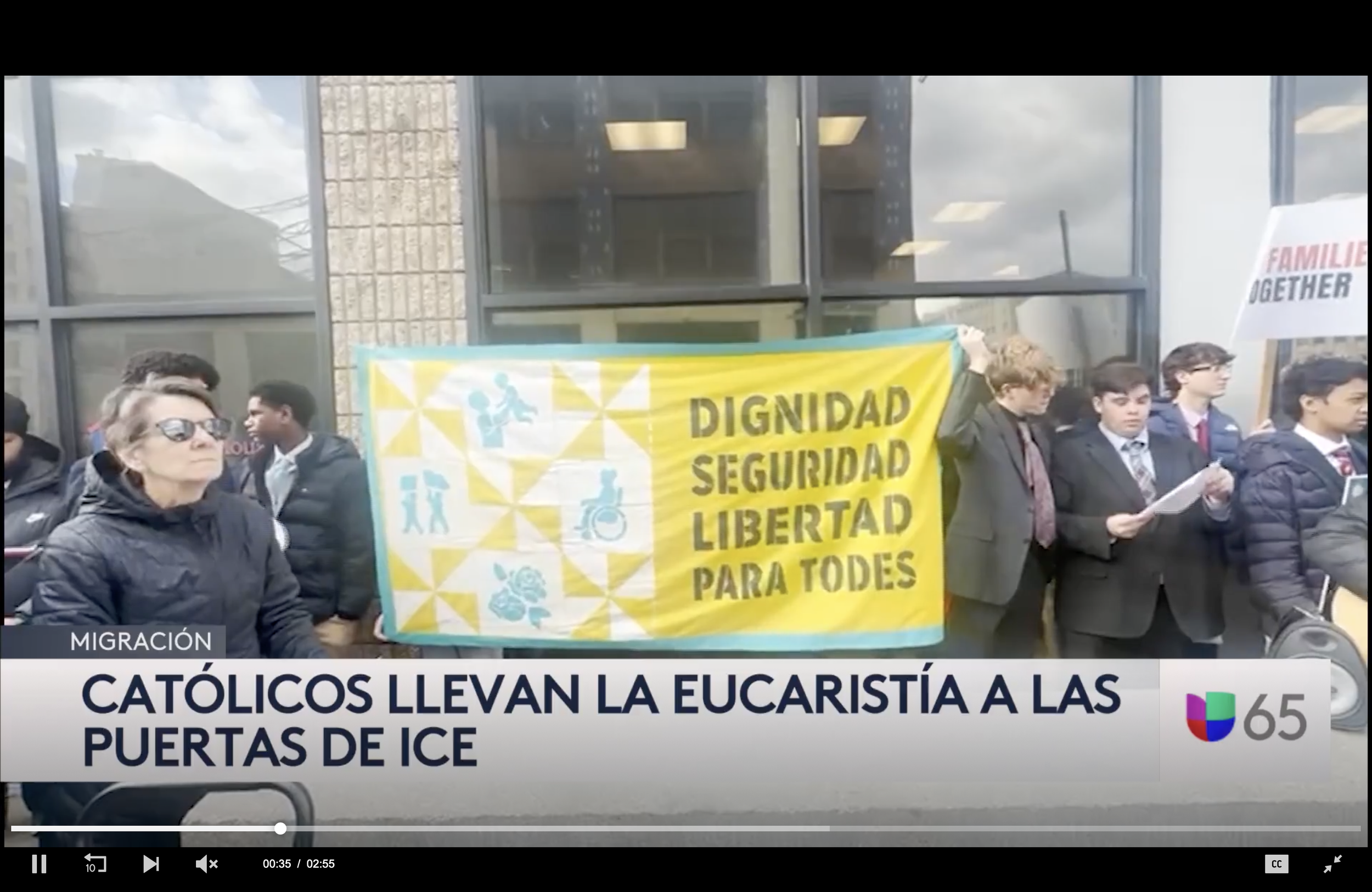 A group of people participating in a protest march outside a building, holding a yellow banner with social and disability icons and words in Spanish about dignity, security, and freedom for all. The news caption in Spanish reads, 'Catholics bring the Eucharist to ICE doors.'