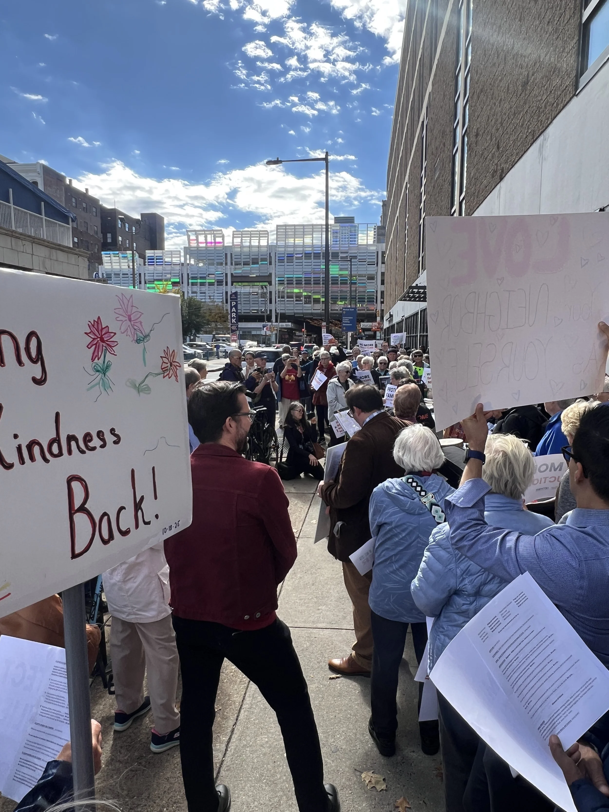 Crowd of people protesting in front of the Philadelphia ICE field office holding signs with messages about kindness and love, some reading printed papers. The sky is bright with scattered clouds and tall buildings are visible in the background.
