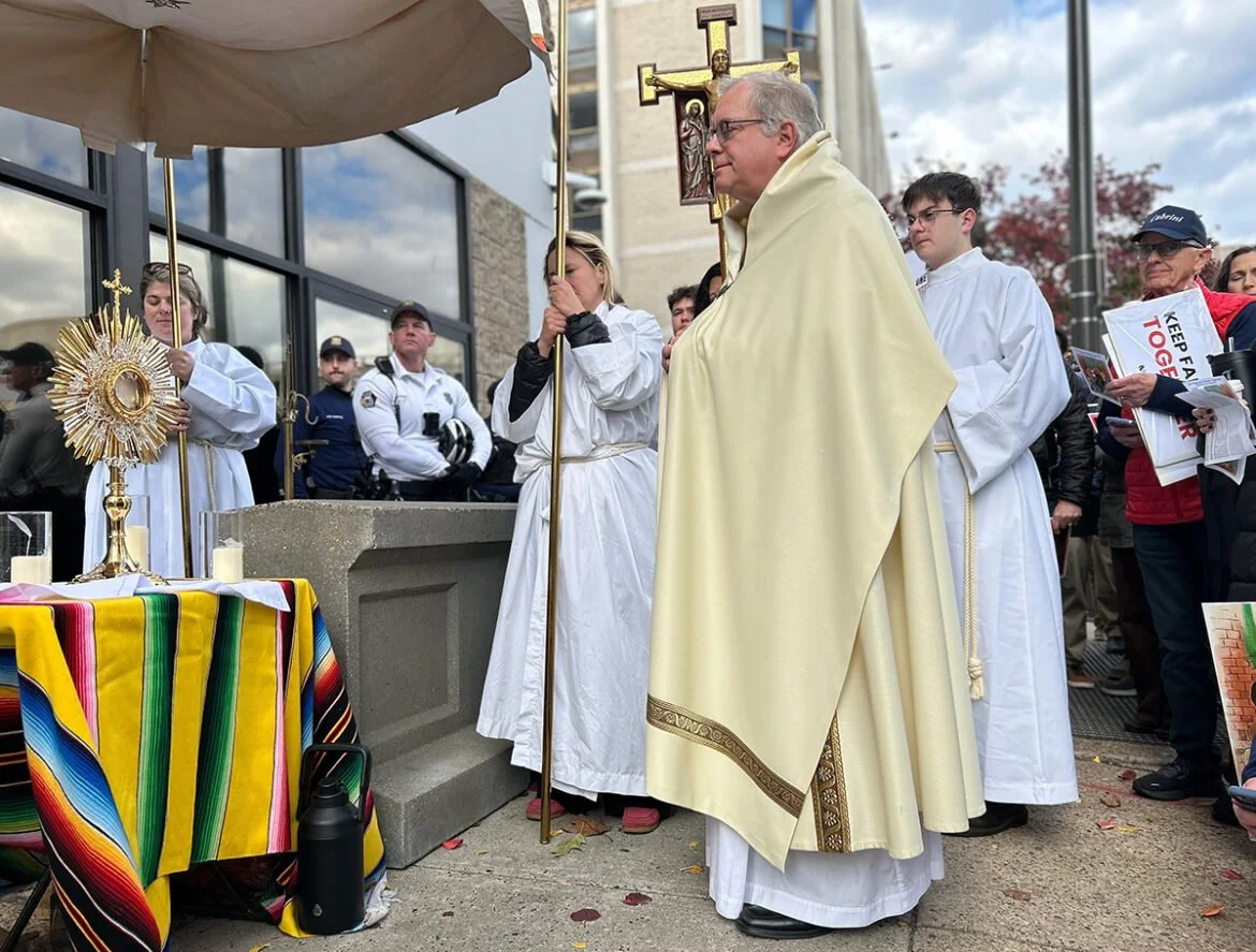 A priest dressed in yellow robes leading a religious procession outdoors, surrounded by altar servers in white robes, with a table covered in a multicolored cloth holding a monstrance, candles, and a chalice, while police officers and onlookers are visible in the background.