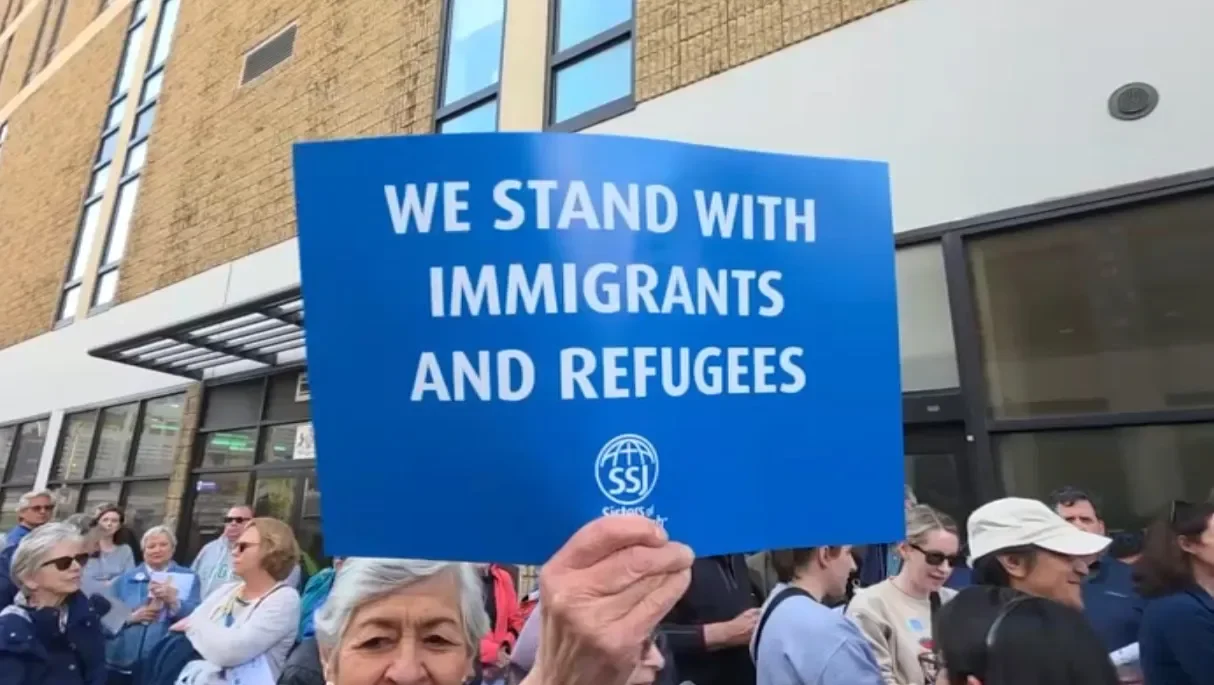 A woman holding a blue sign that reads 'We stand with immigrants and refugees' at a rally or protest with a crowd of diverse people in the background.