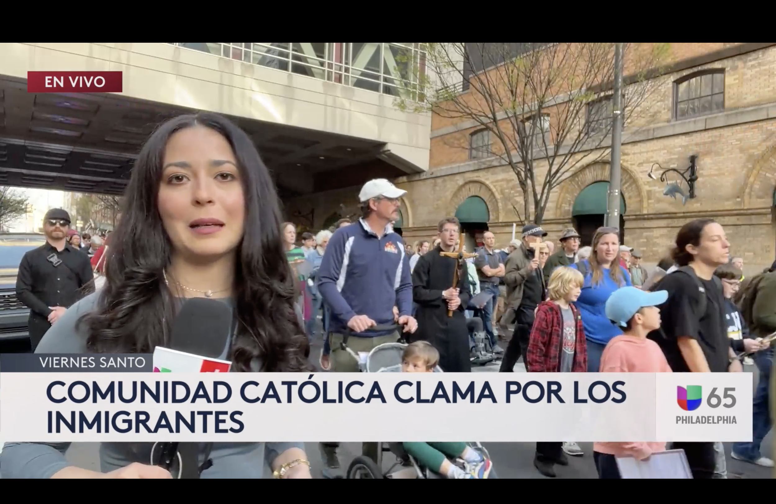 News reporter in front of a crowd during a protest in Philadelphia, holding a microphone with a Barcelona logo, with people of different ages and backgrounds gathered around, demonstrating, and some holding signs.