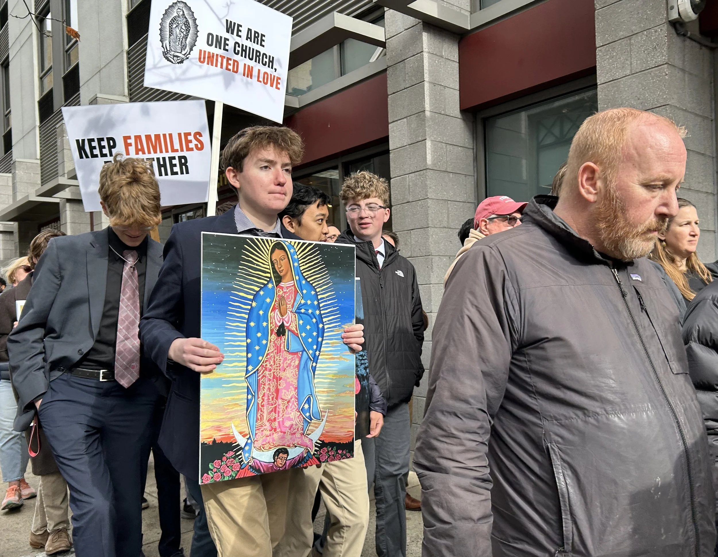 A group of people at a march or rally, some holding signs with messages like 'We are one church, united in love' and 'Keep families together.' One young man in the center holds a picture of Our Lady of Guadalupe, a religious icon.