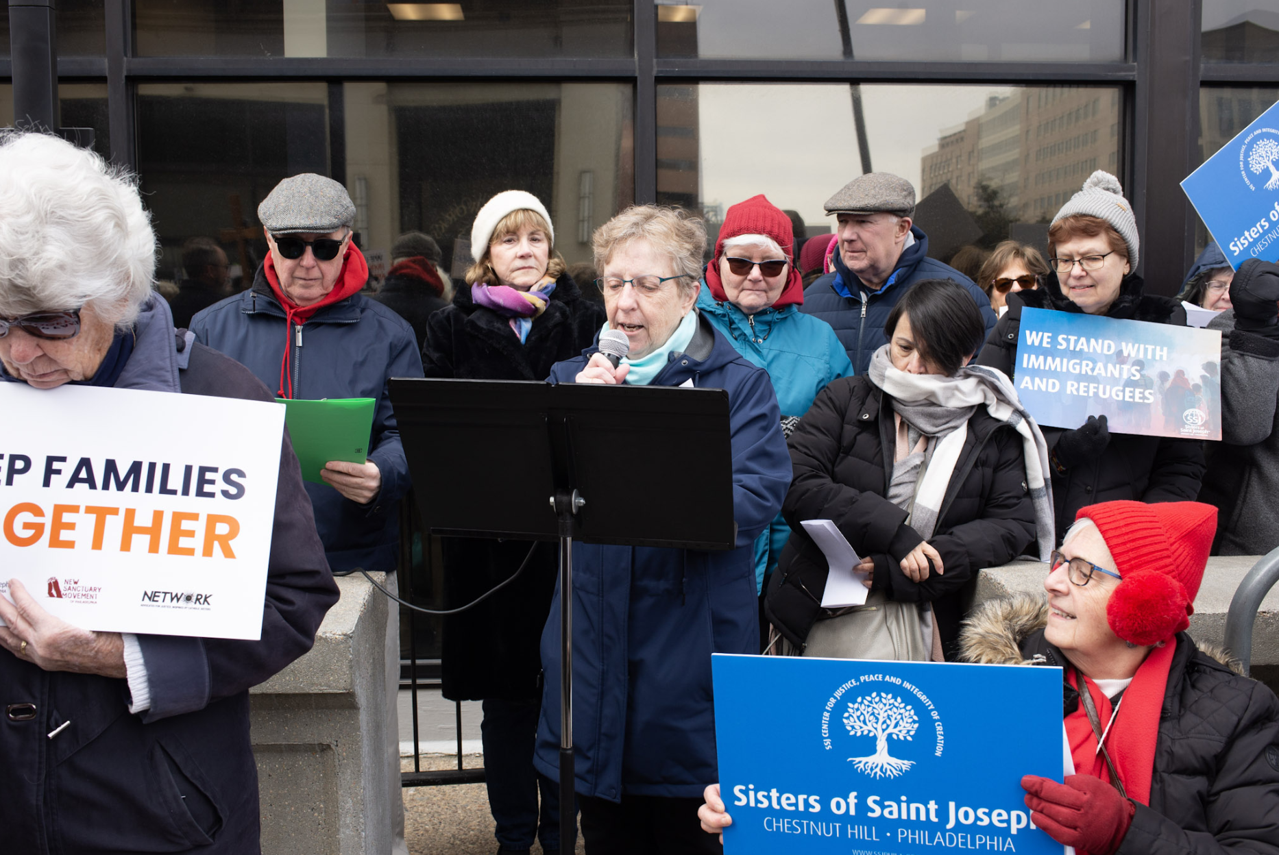 A group of people participating in a demonstration or rally outside a building. A woman stands at a microphone reading, surrounded by others holding signs, including one that says "We stand with immigrants and refugees" and another with the name "Sisters of Saint Joseph, Chestnut Hill, Philadelphia." Most of the participants are dressed warmly in winter clothing, with some wearing hats and scarves.